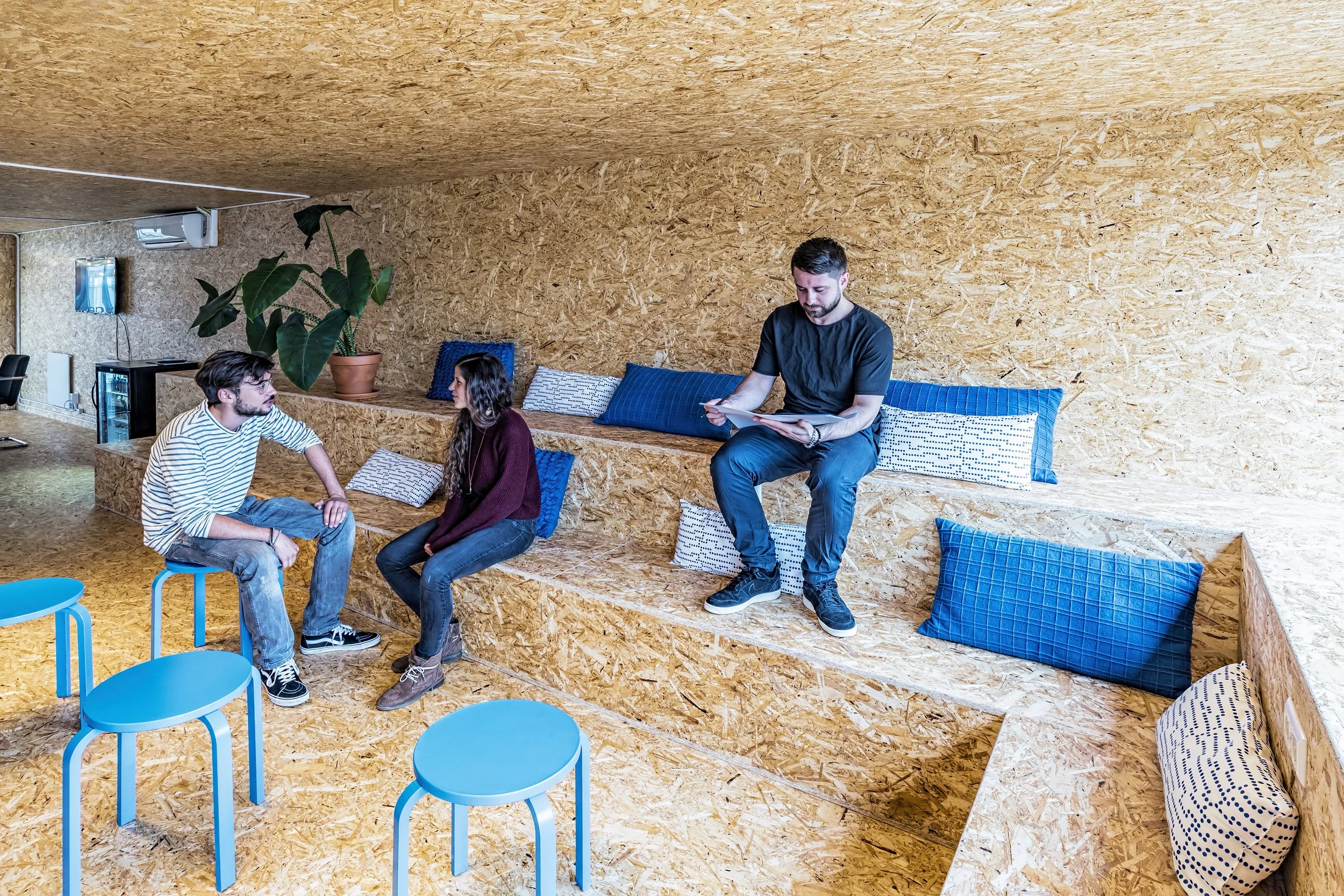 Three people sitting on wooden benches with blue cushions inside a room with wooden walls, two men and one woman, one man holding a tablet, the other man wearing a striped shirt, and the woman with long hair in a maroon sweater.