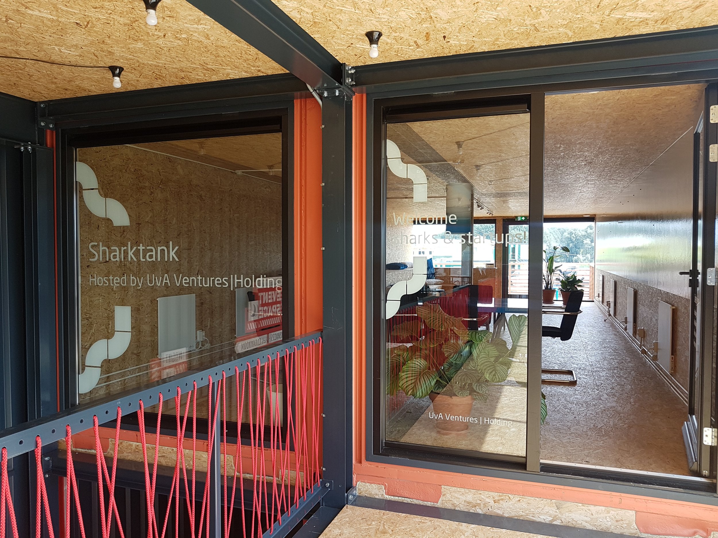 Interior view of an office space with wooden ceiling, glass walls, and a welcome sign that reads 'Welcome Sharks & Starups! Hosted by UVA Ventures Holding.' There is a potted plant and a seating area visible through the glass.