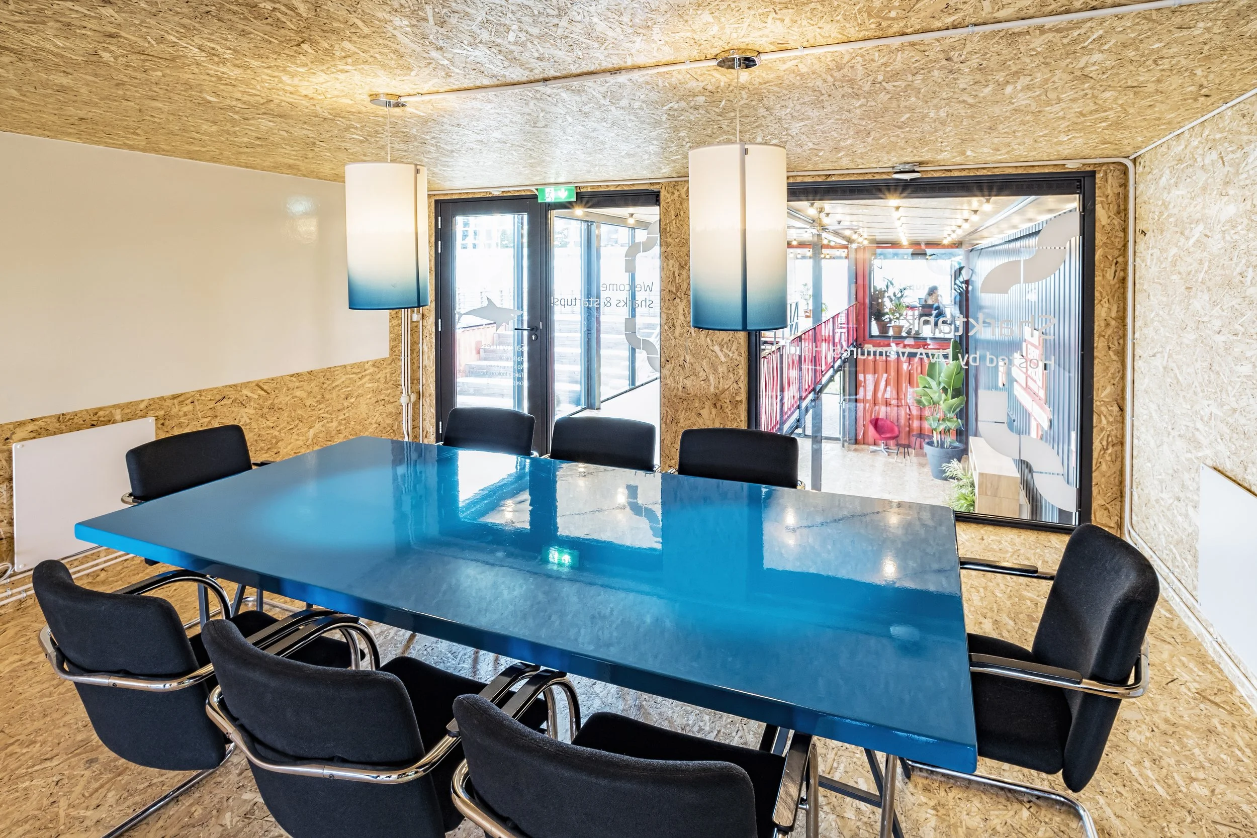 A modern conference room with a bright blue table, black chairs, wood-textured walls and ceiling, large glass doors, and hanging blue pendant lights.