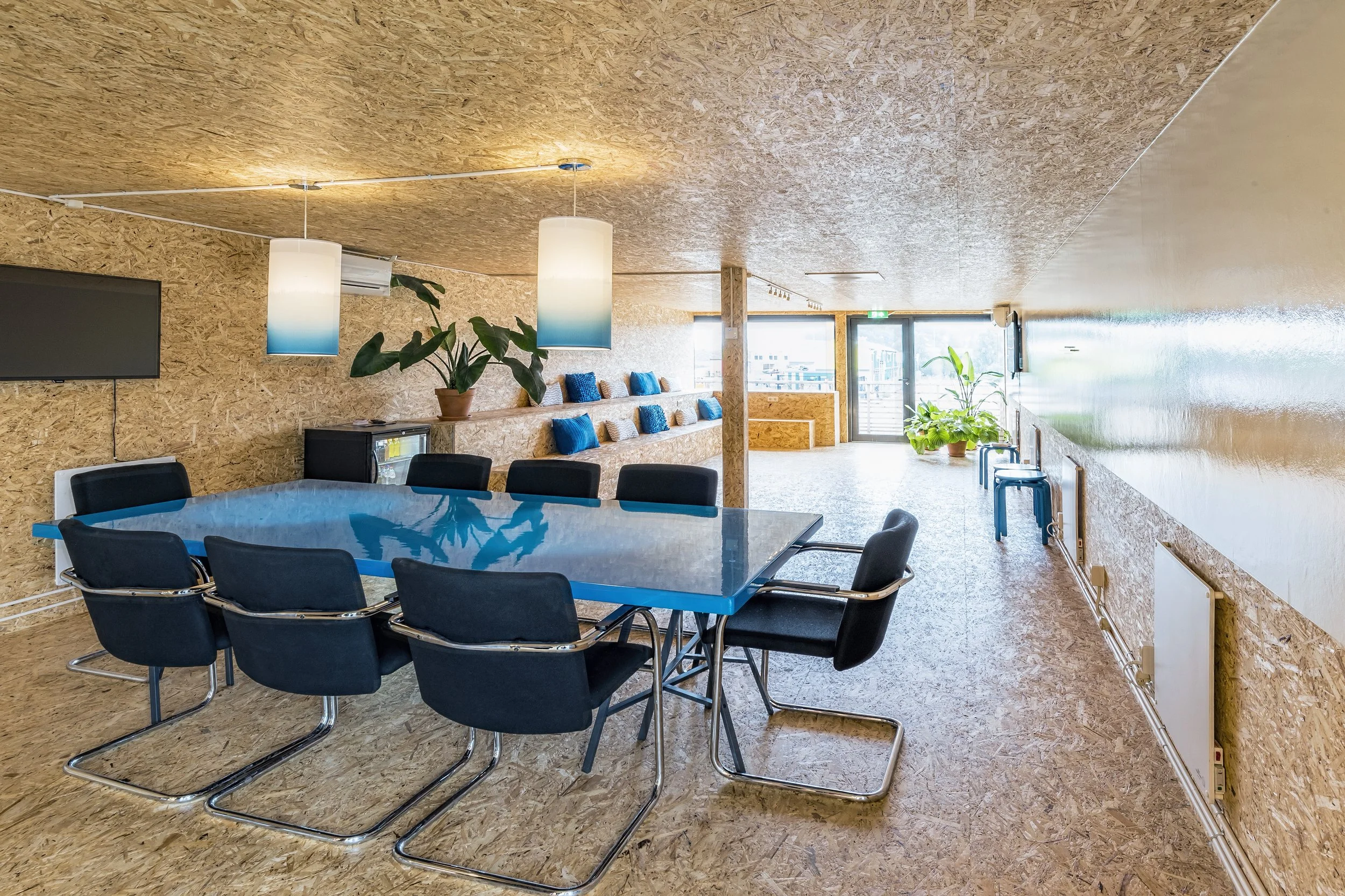 Modern conference room with a large blue table, black chairs, potted plants, pendant lights, and large windows providing natural light.
