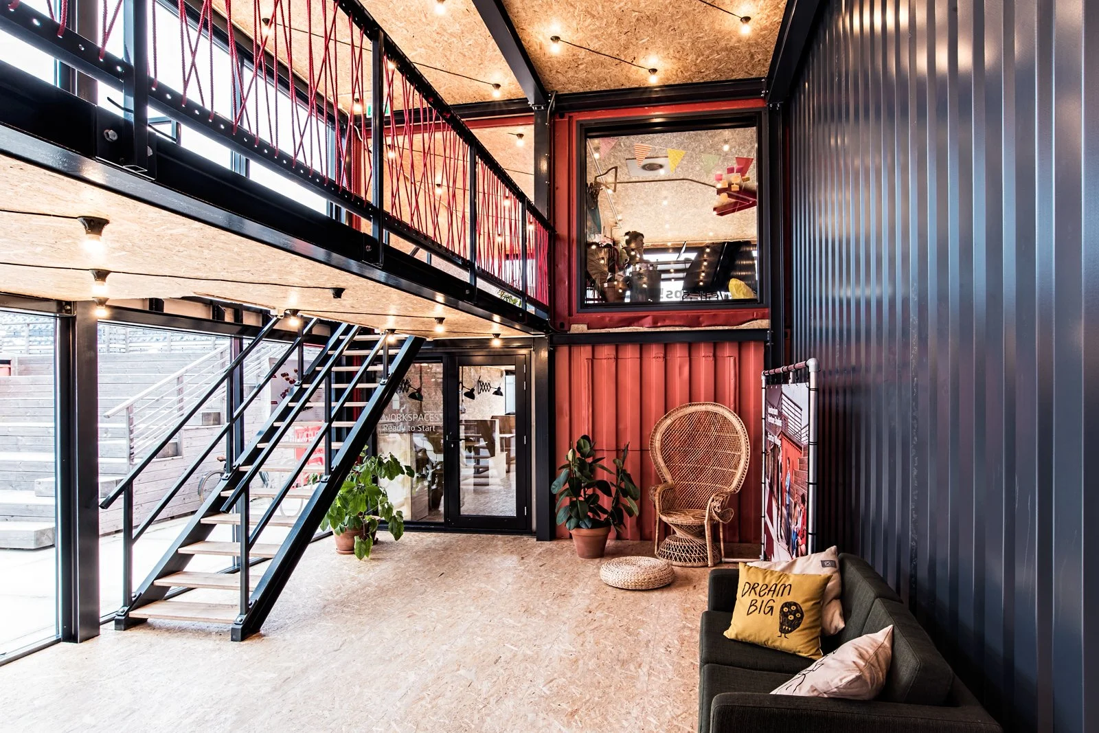 Interior of a modern, industrial-style space with a black metal staircase, potted plants, wicker chair, decorative pillows, and a glass-enclosed room upstairs, illuminated by string lights.