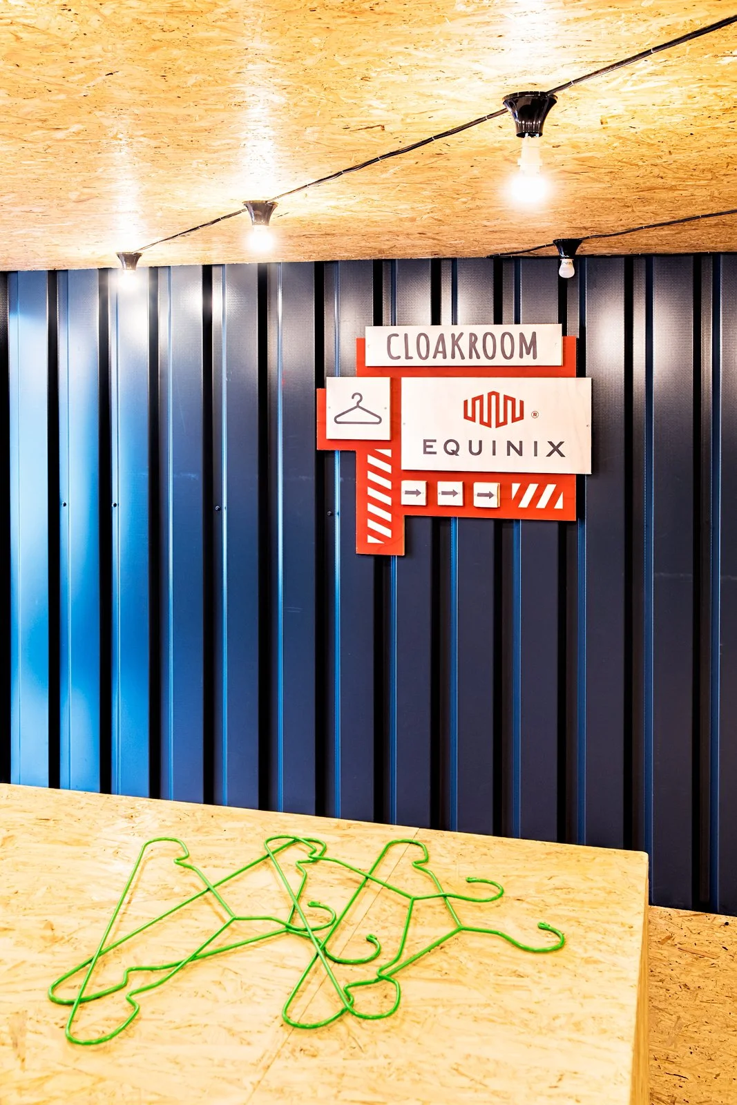 Sign with words 'Cloakroom' and 'Equinix', a clothing hanger icon, and directional arrows, mounted on a dark blue corrugated wall. Bright lights are hanging from a wooden ceiling, and a wooden table in the foreground has several green wire hangers.