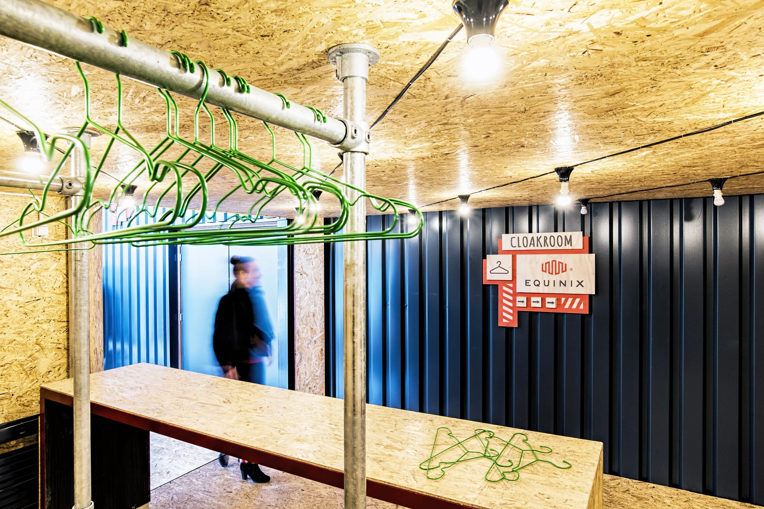 Interior of a retail space with a wooden ceiling, green hangers on a metal rod, and a sign that reads 'Cloakroom' with arrows, black corrugated panel wall, and a person blurred in motion.