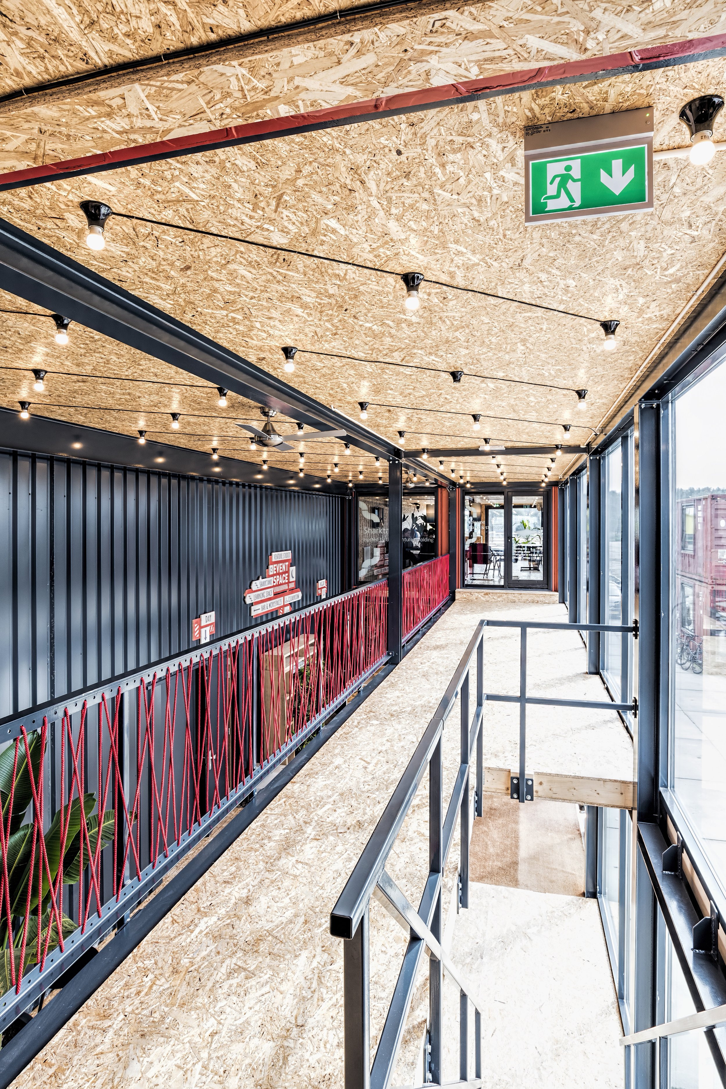 Interior of a modern building with a wooden ceiling, string lights, glass walls, and a ramp with metal railing leading to a door. There is a green exit sign with a running figure and downward arrow on the ceiling.