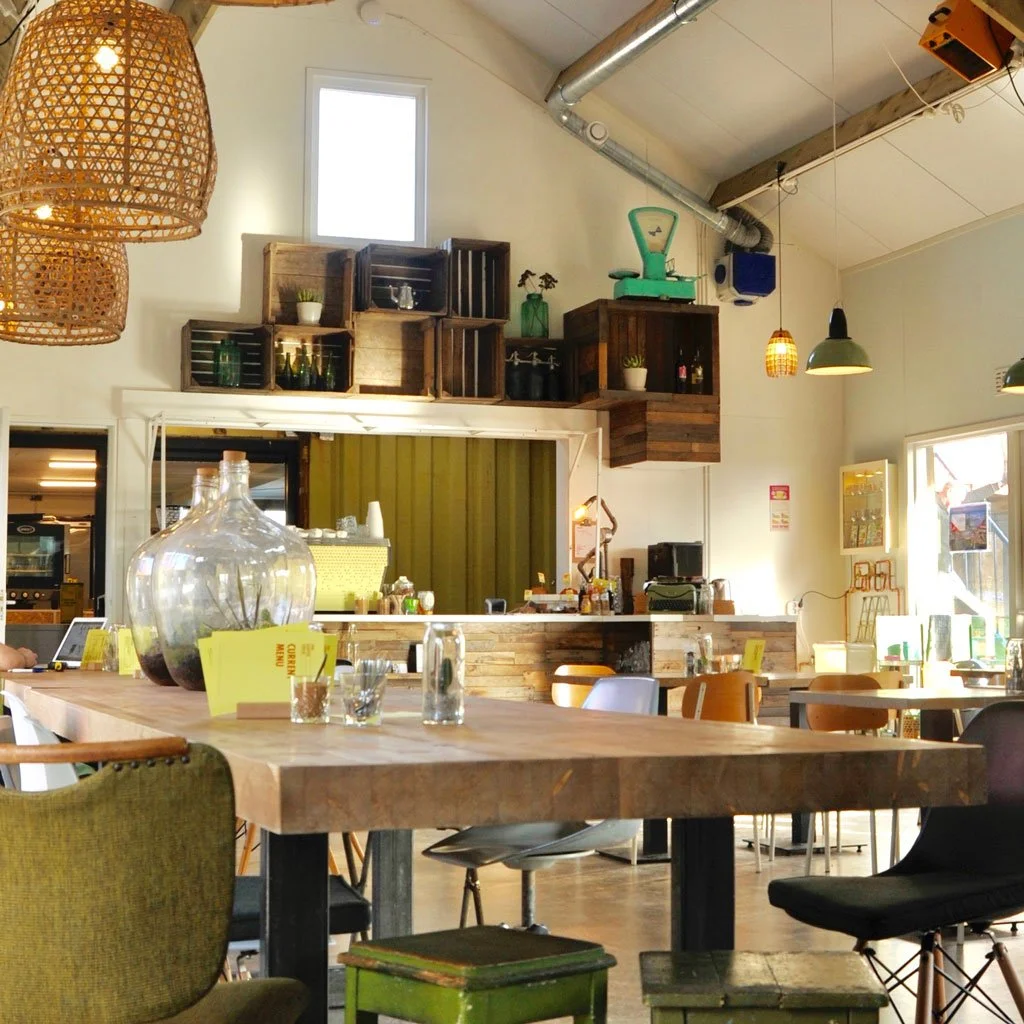 Inside view of a cozy cafe with wooden tables and various colorful chairs, decorative wooden crates and bottles on a high shelf, pendant lights, and a large glass jar on the table.