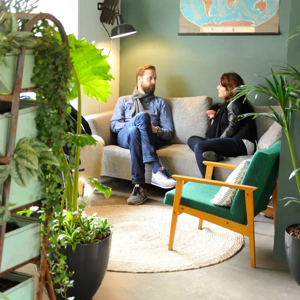 Two people sitting on a beige sofa having a conversation in a cozy, green-walled room decorated with plants and a map on the wall.