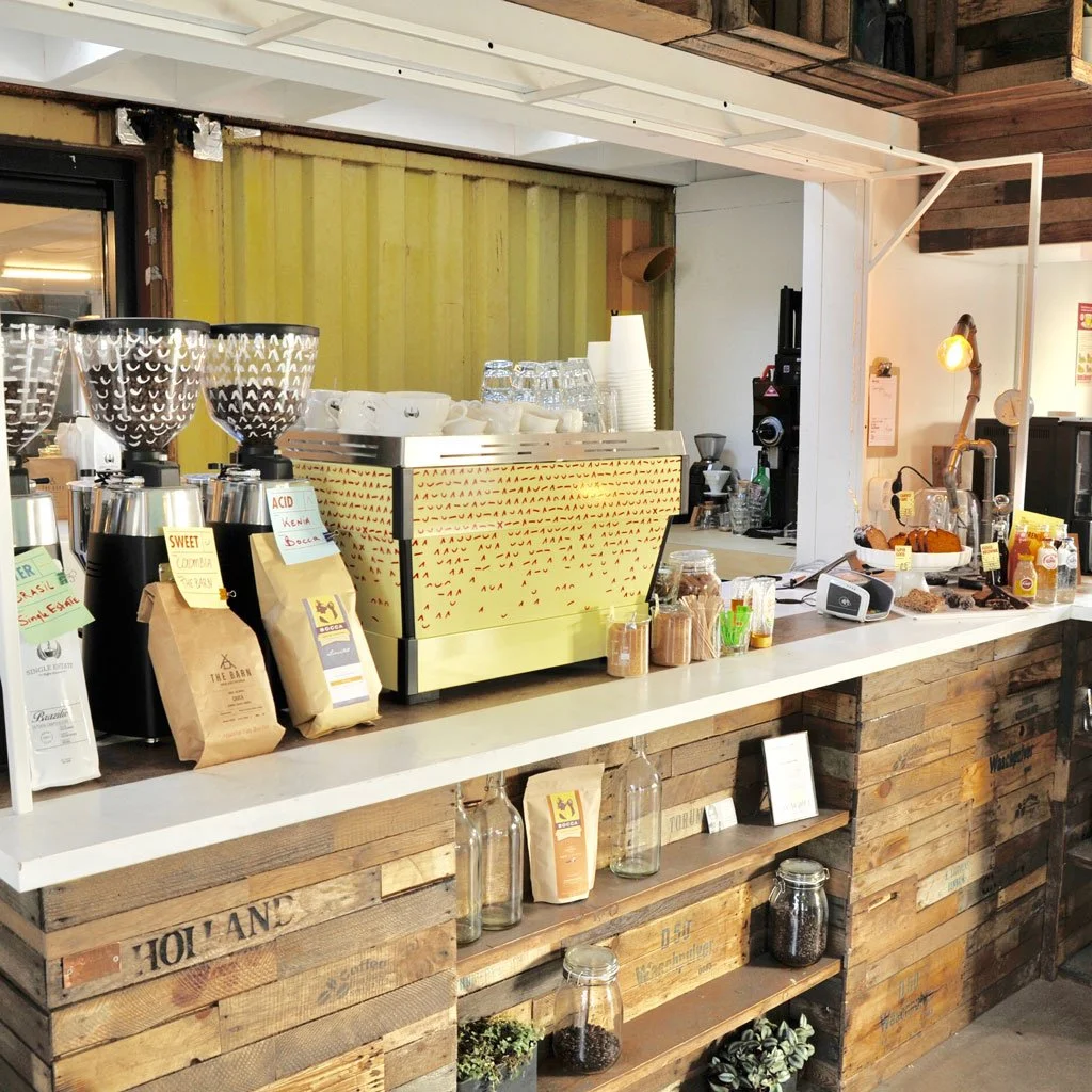 Coffee station with a yellow front and various coffee beans, cups, and brewing supplies on a white counter, in a rustic cafe setting.