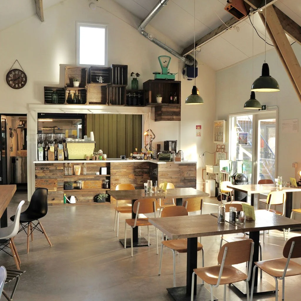 Interior of a coffee shop with wooden tables and chairs, a counter with coffee equipment, and large hanging lights.
