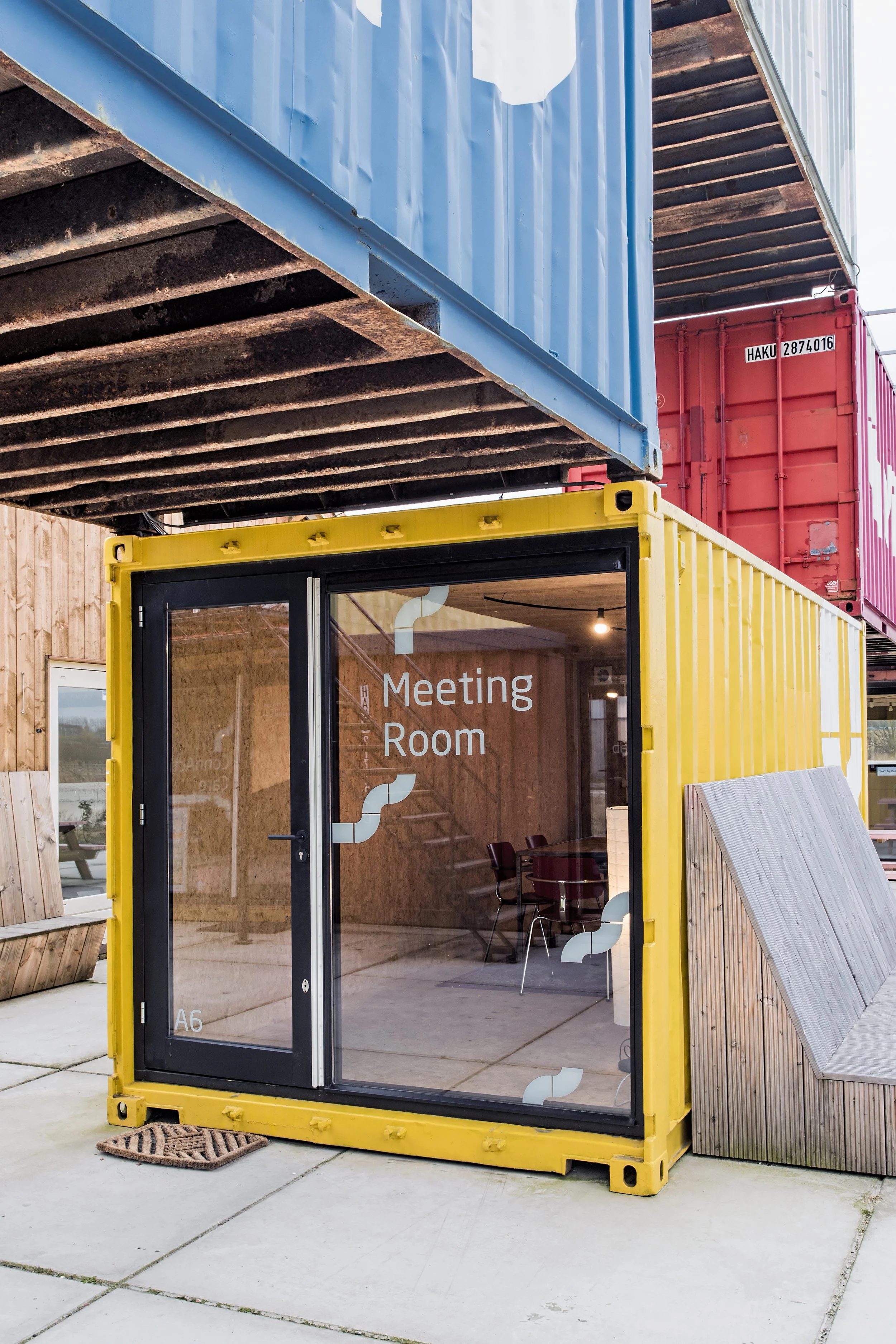 A yellow container with glass walls serving as a meeting room, positioned outdoors on a concrete patio with a doormat in front. Stacked shipping containers are visible above and behind it.