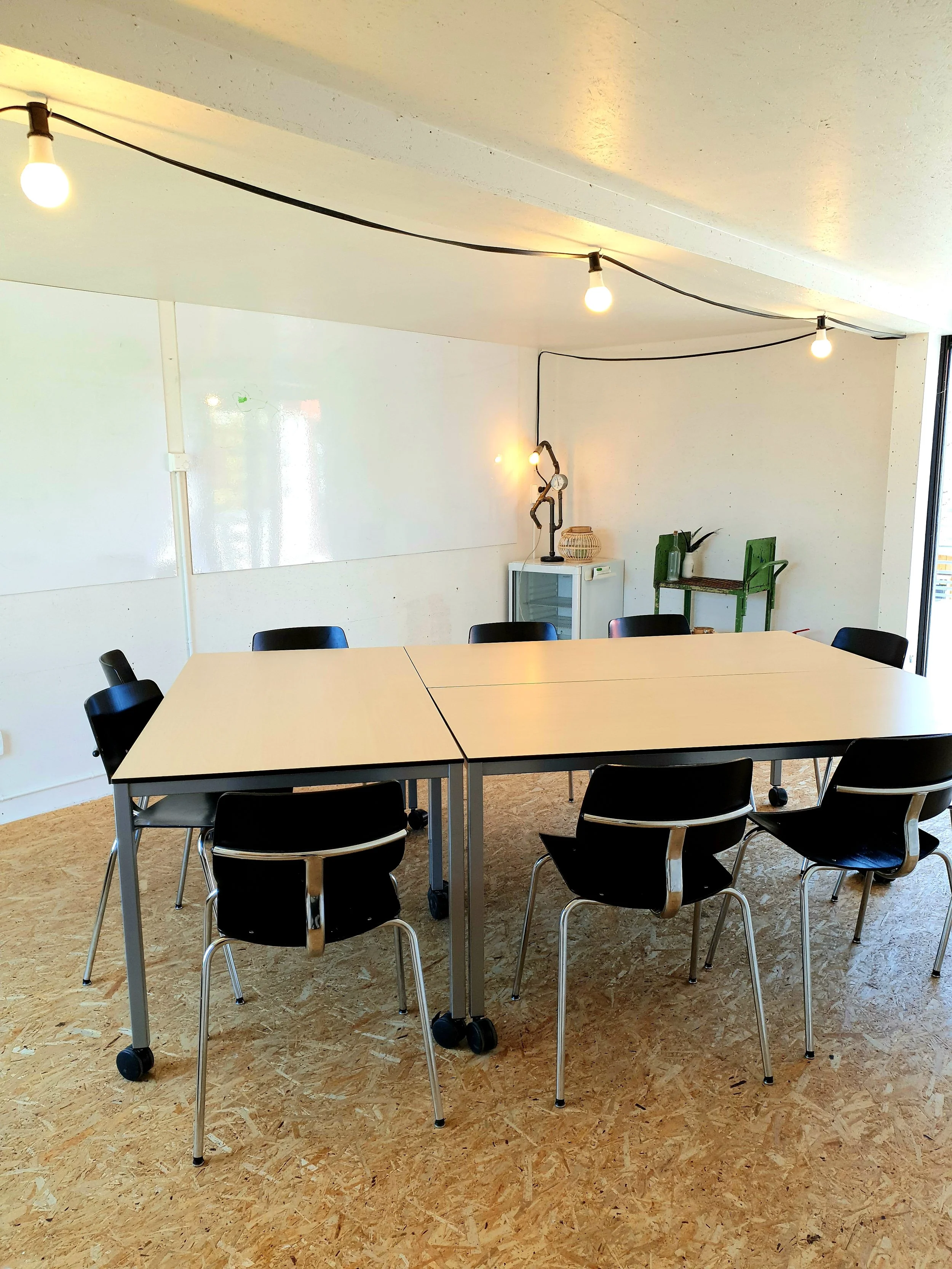 A room with a long table surrounded by black chairs, a white wall with a whiteboard, a small cabinet, and green chairs stacked with vases and long plant stems. Overhead lights are strung across the ceiling.
