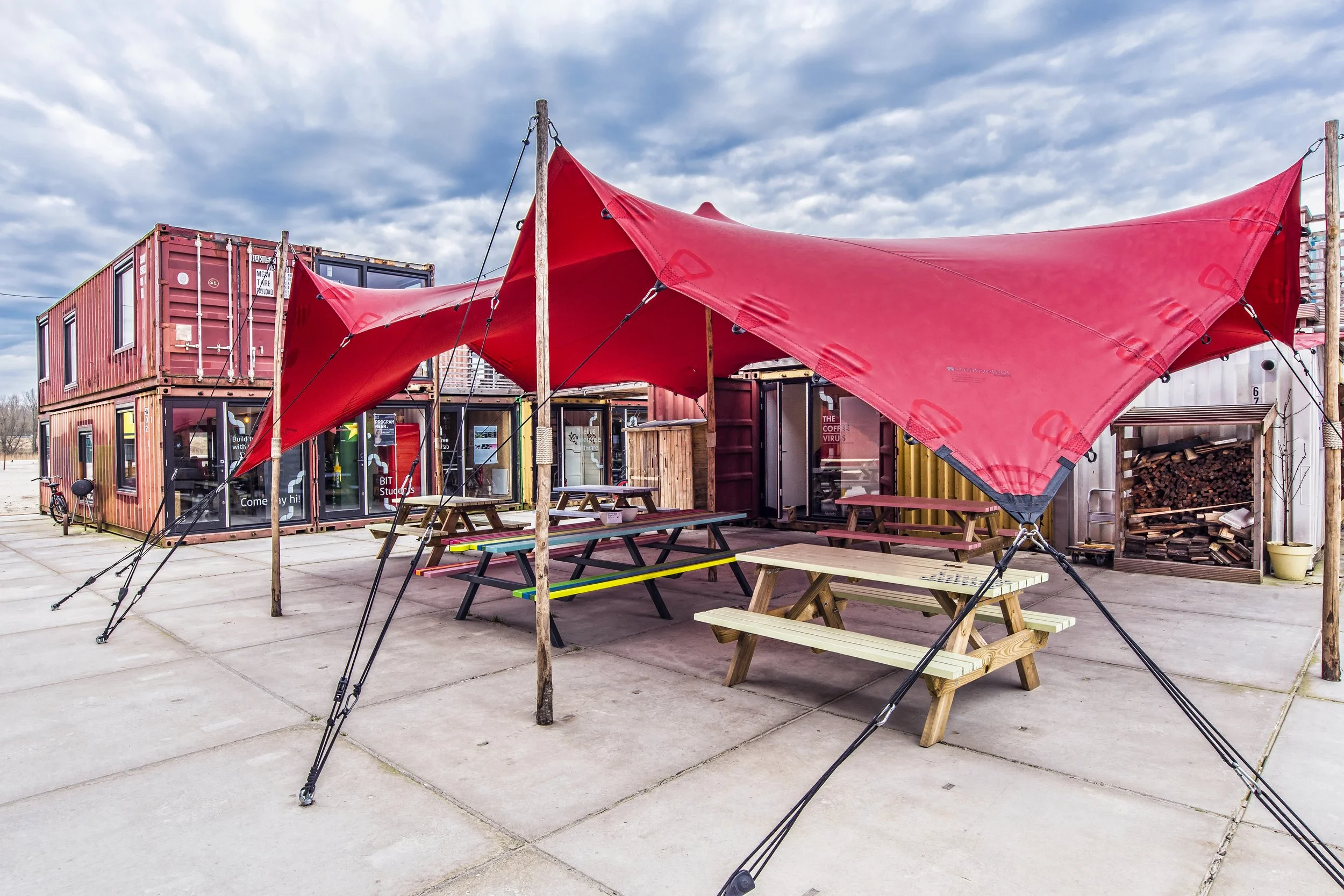 A courtyard with shipping containers repurposed as buildings, featuring picnic tables and a large red fabric canopy supported by wooden poles, with a cloudy sky overhead.