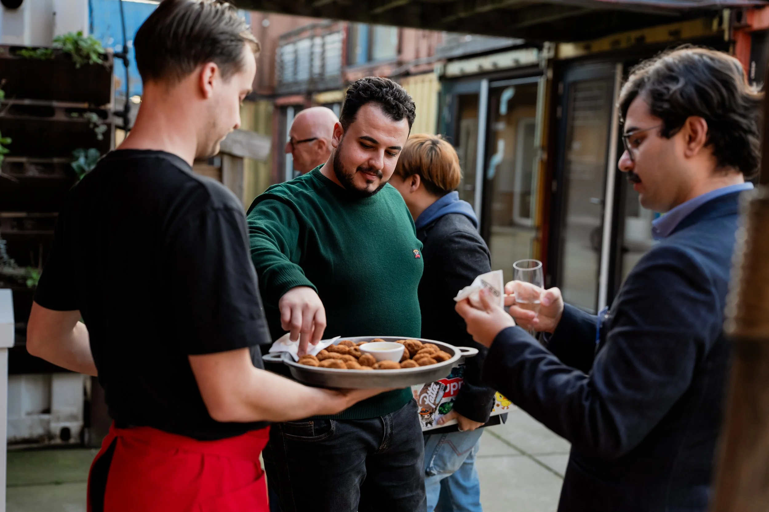 People gathered outdoors at a social event, with one person serving food, specifically a tray of fried food with dipping sauce, while others reach for it and interact.