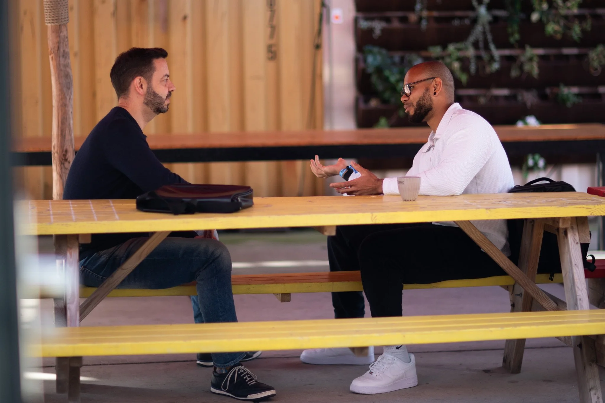 Two men sitting at a wooden picnic table engaged in conversation, one gesturing with his hand, with greenery and a wooden fence in the background.