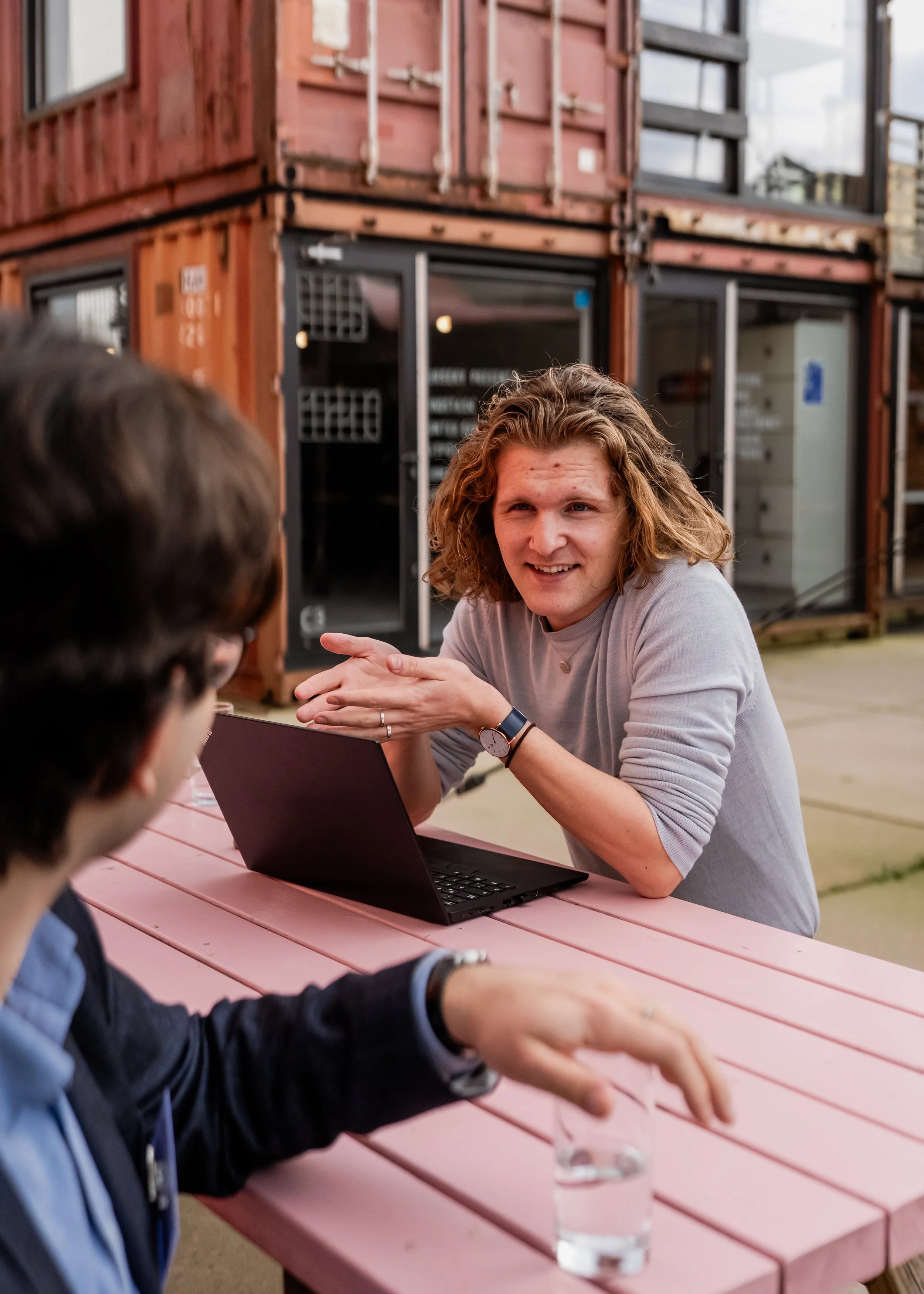 A woman with shoulder-length blond hair and a gray shirt is sitting at a pink table, smiling and gesturing with her hands while talking to a group of people outdoors during daytime. There is a laptop and a glass of water in front of her, and a building with a modern design made of shipping containers is visible in the background.