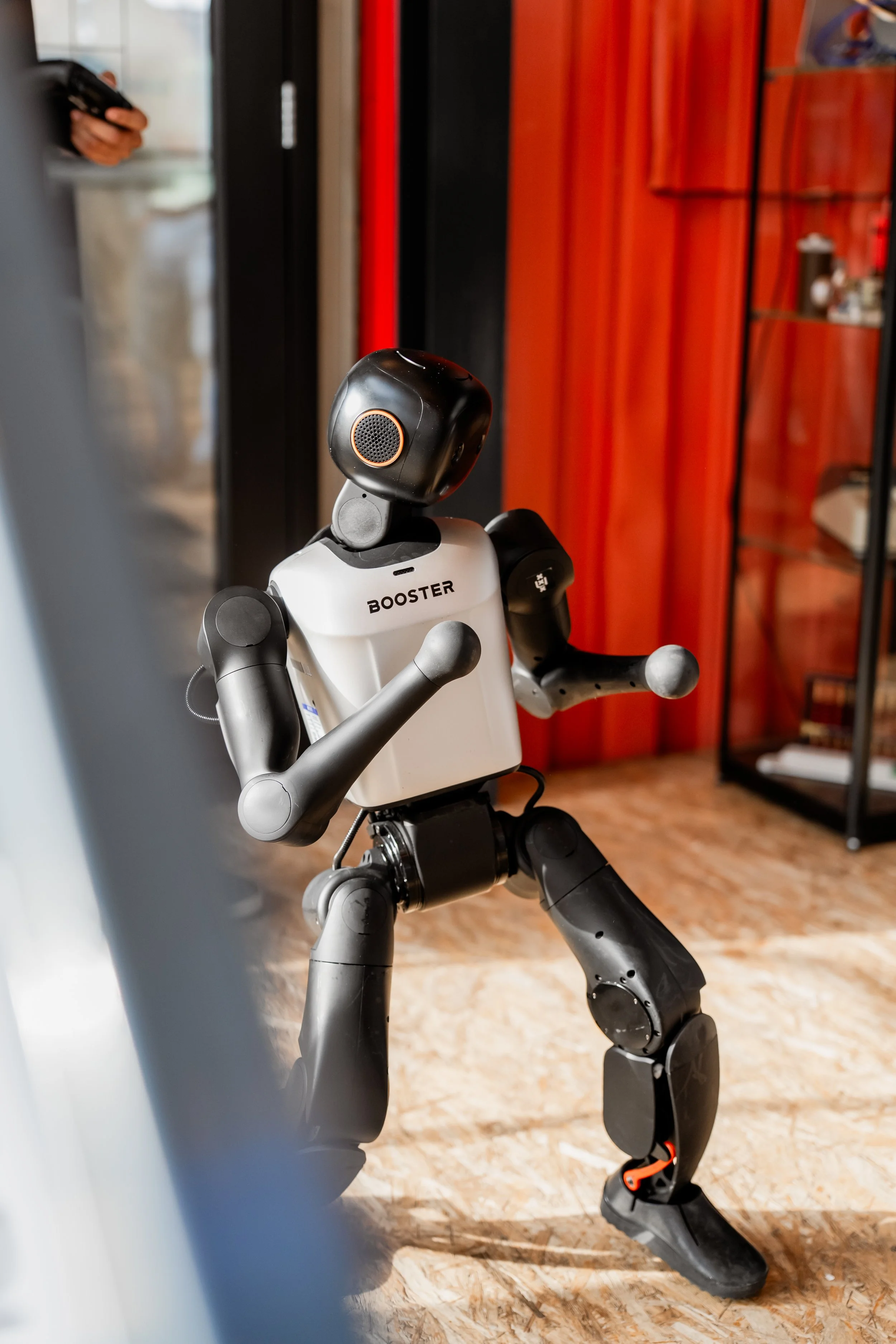 A small humanoid robot with a white torso labeled "BOOSTER" standing on a hardwood floor. The robot has black arms and legs, with orange accents, and an oval-shaped head with a speaker grille in orange. In the background, there's a red and black bookshelf and a person holding a remote control.