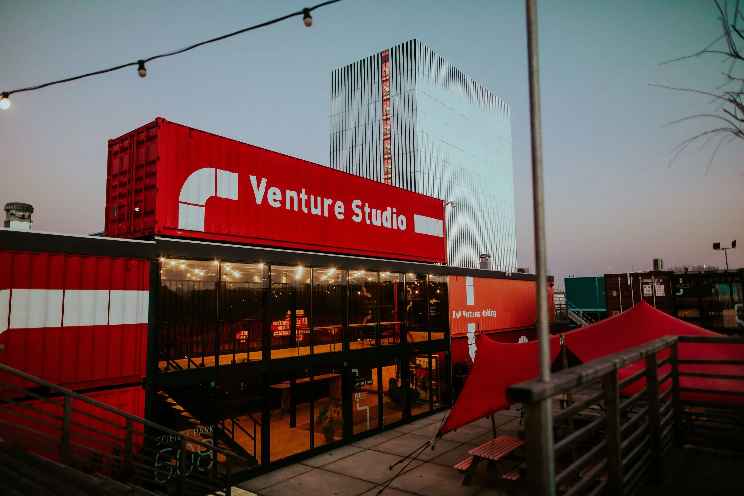 View of a modern building with a large red sign reading "Venture Studio". The building has a glass facade with visible interior lights and is constructed from shipping containers painted red and white. There are string lights and an outdoor seating area with picnic tables and benches, set against a twilight sky.