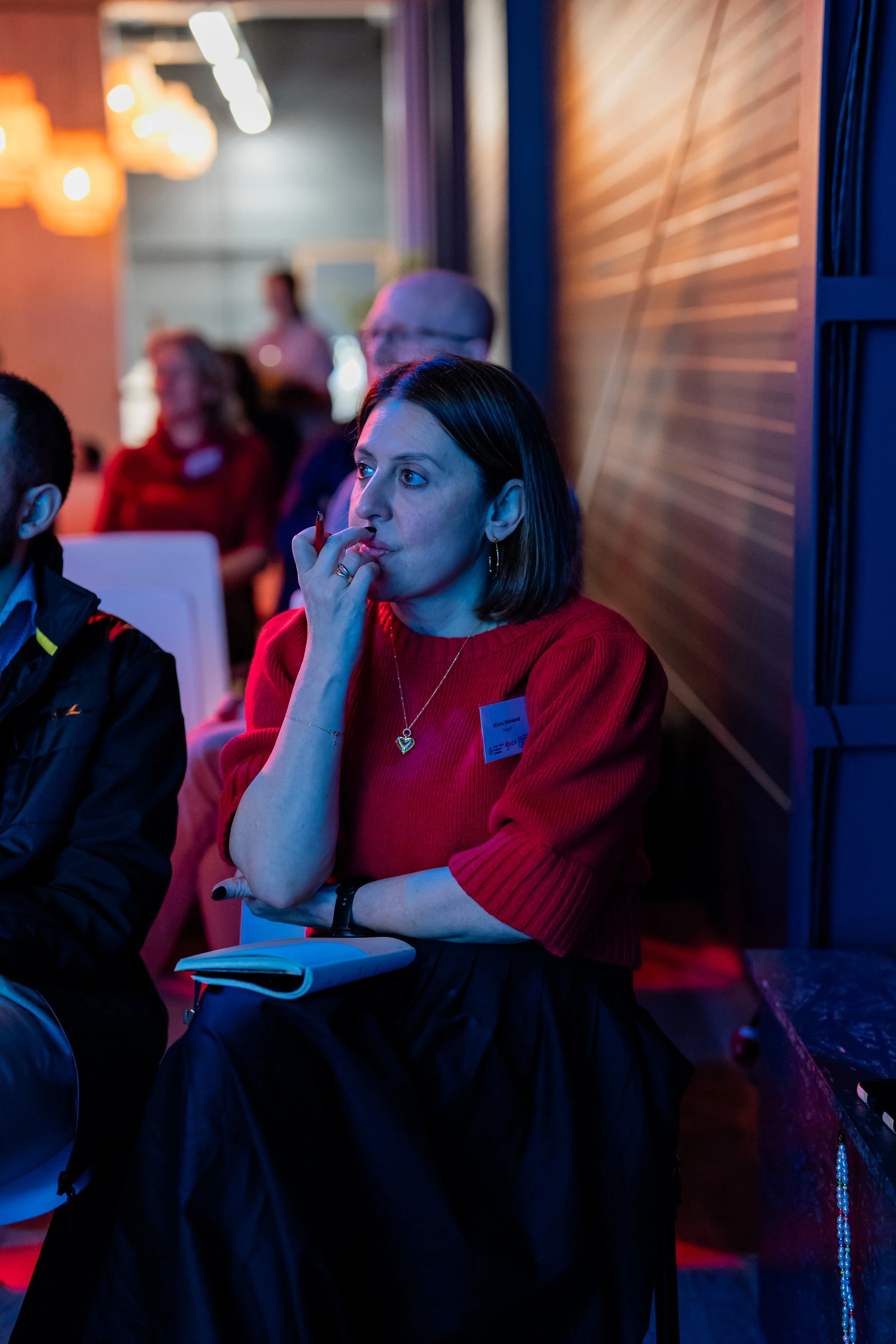 A woman with short dark hair wearing a red sweater and gold hoop earrings, sitting at an indoor event, holding a notebook, with her hand near her mouth, listening attentively.