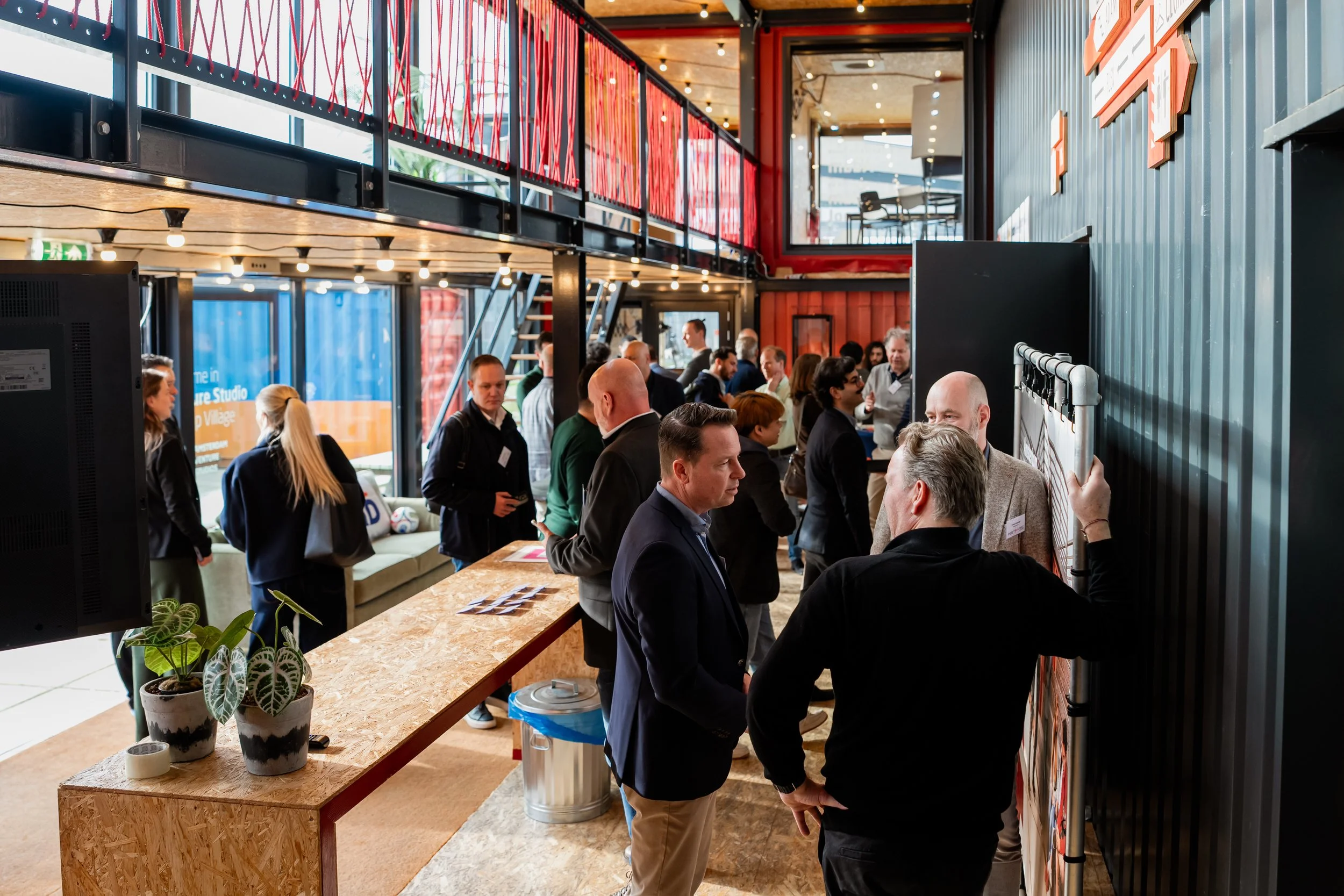 A crowded indoor event with people engaging in conversations, a table with potted plants, and a high ceiling with exposed wood and lighting fixtures. There are red and black accents and large windows in the background.