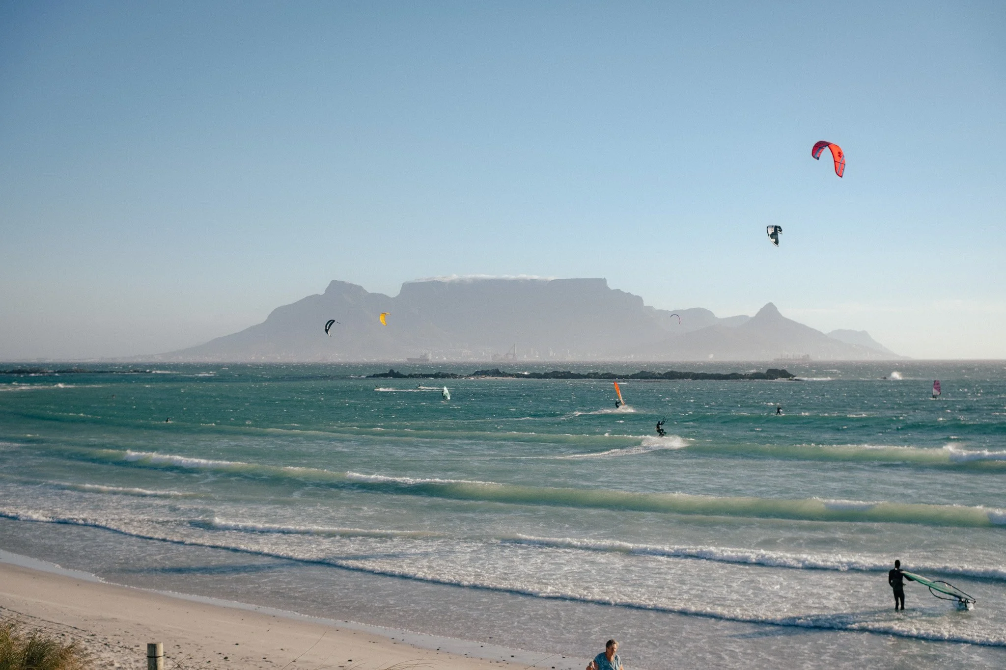 Kite surfers riding the waves in the ocean near a sandy beach, with mountains visible in the background under a clear sky.
