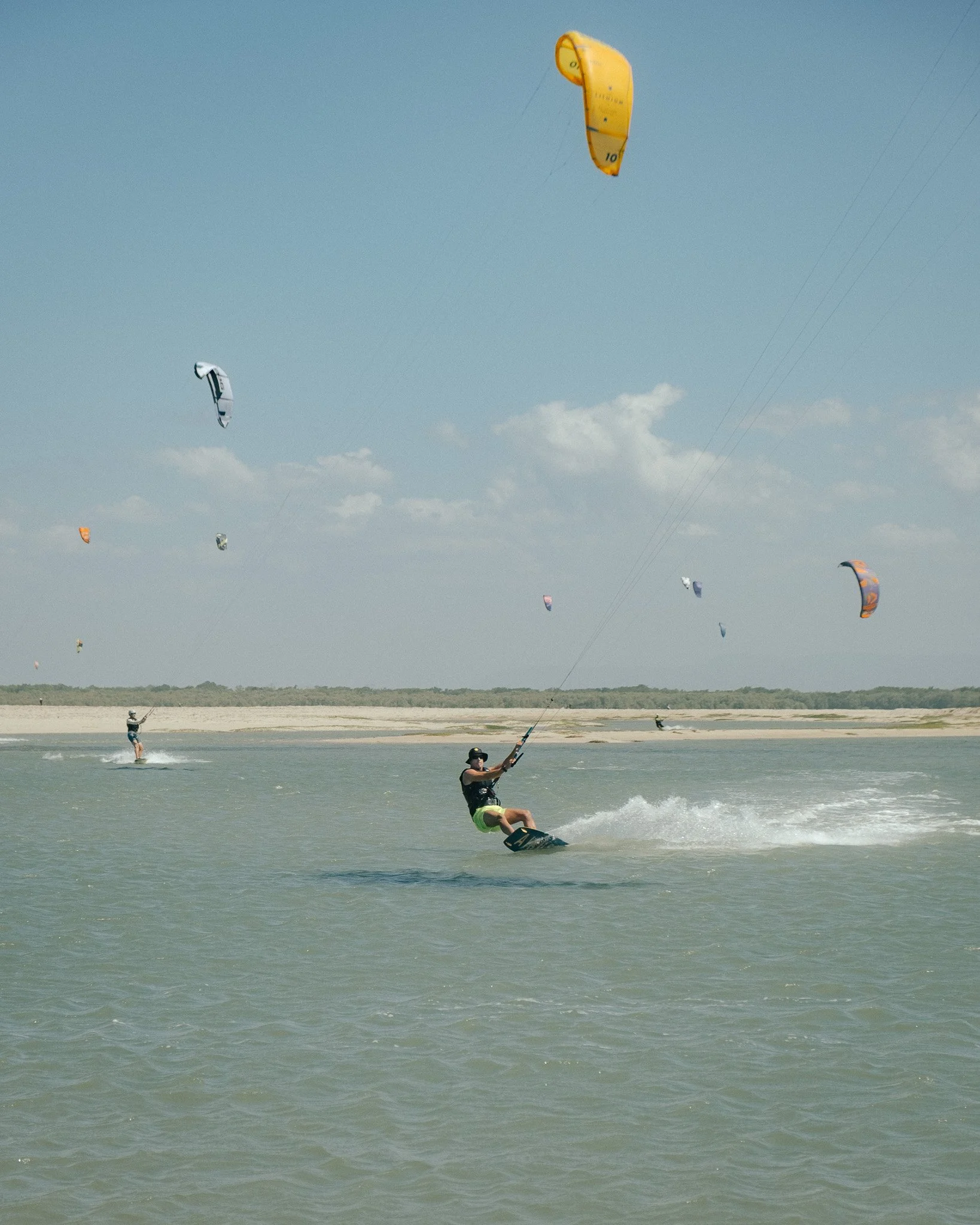 Multiple people kiteboarding on the ocean, with colorful kites in the sky and some people in the background on the beach.