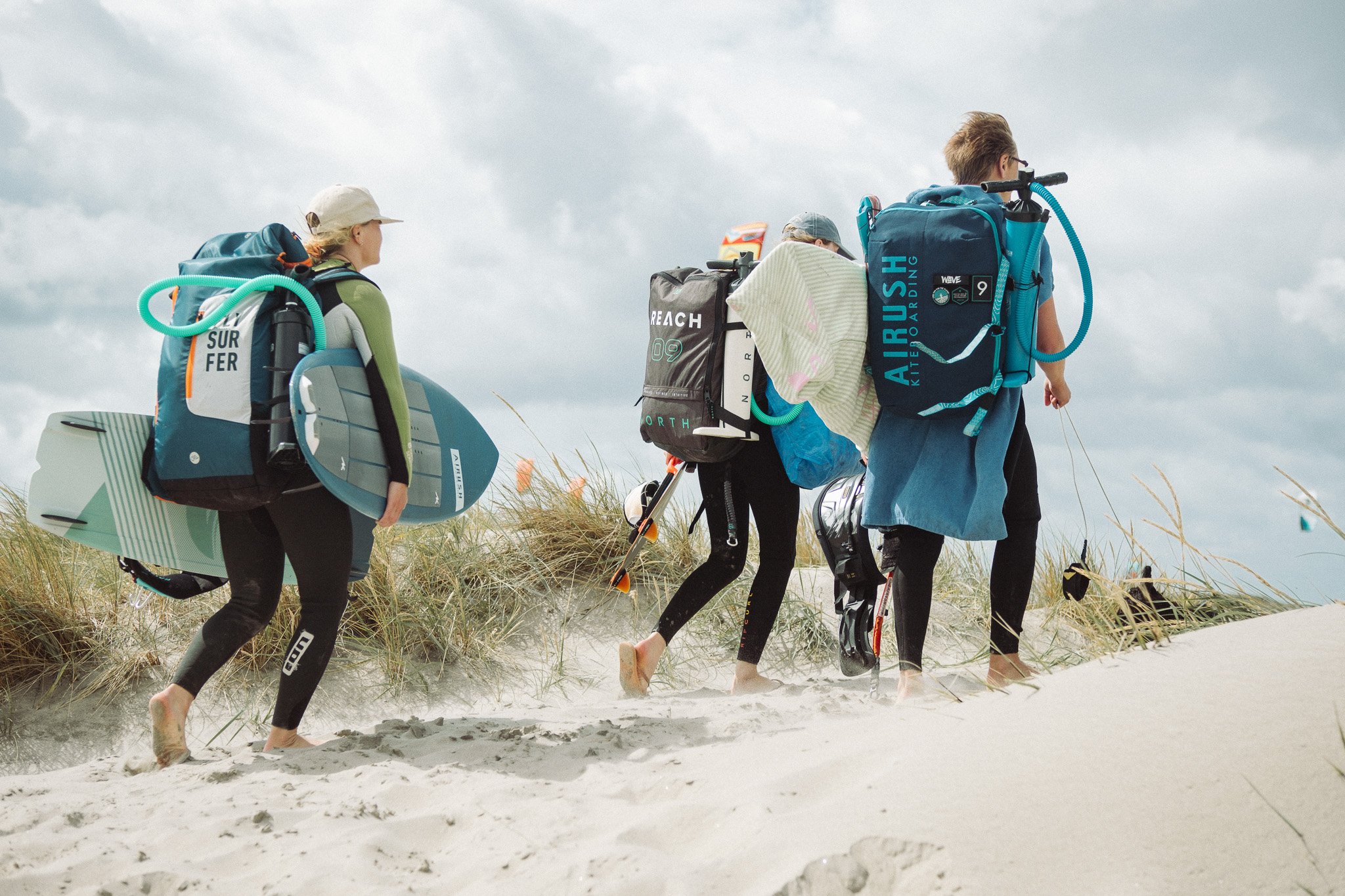 Group of children walking barefoot on a sandy beach, carrying surfboards and backpacks, with cloudy sky overhead.