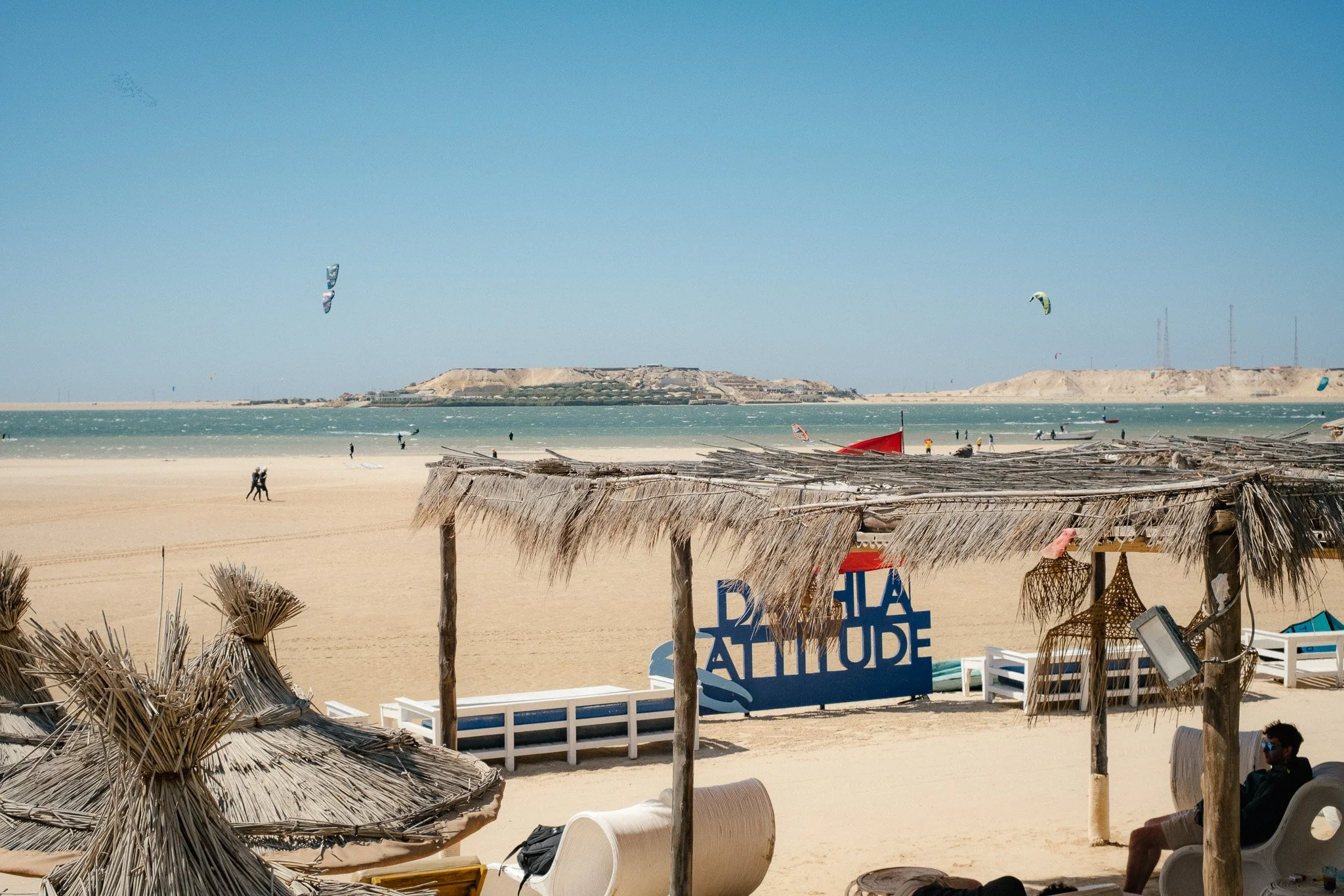Beach scene with thatched umbrellas, lounge chairs, and a sign that reads 'Bahia de Atamu' in front of water. Kitesurfers and people are visible, with a sandy hill in the background and a clear blue sky.