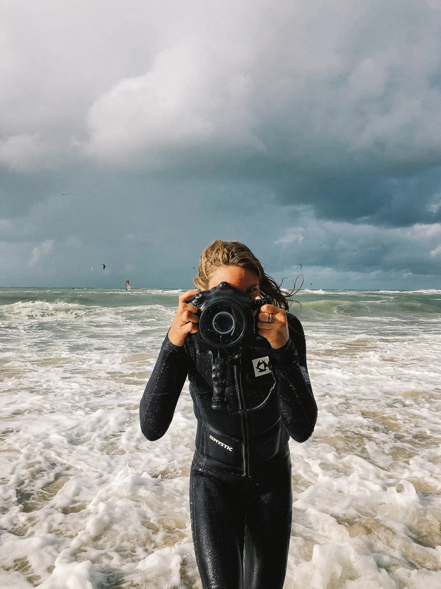 A woman in a wetsuit taking a photo on a beach with cloudy skies and kite surfers in the background.