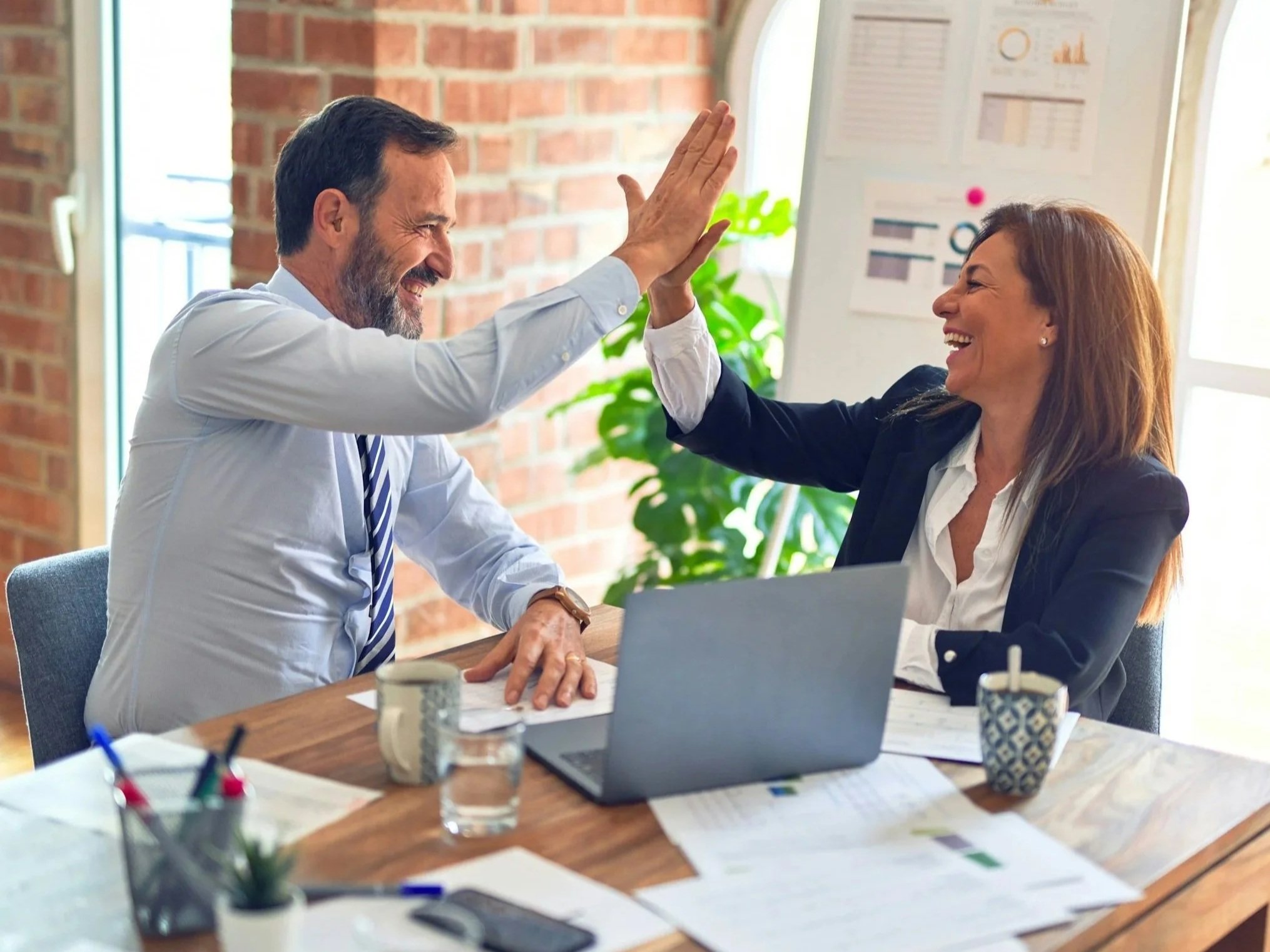 Deux personnes, un homme et une femme, se donnent un high-five en souriant dans une salle de réunion avec un mur en briques. La table entoure des papiers, un ordinateur portable, et des tasse à café, avec un tableau blanc ou une présentation en arrière-plan.