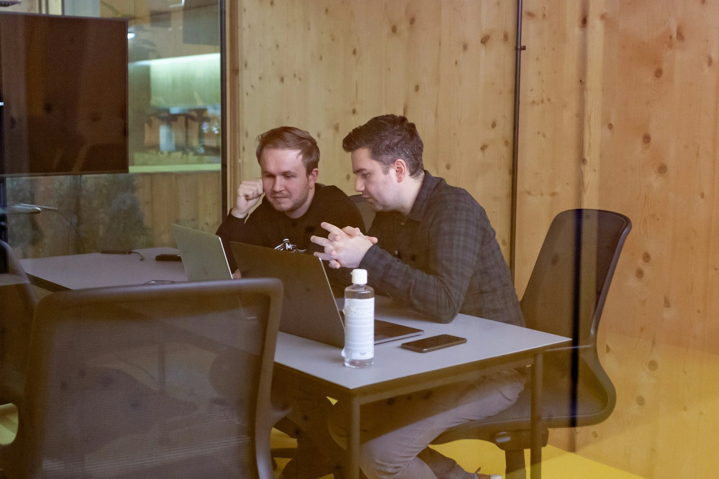 Deux jeunes hommes assis à une table de travail, regardant un ordinateur portable, dans un environnement intérieur en bois.