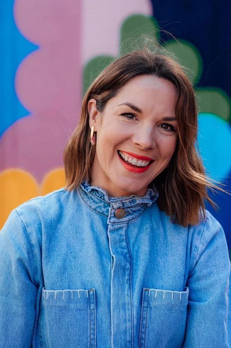 A woman smiling with brown wavy hair, wearing a blue denim jacket and gold hoop earrings, standing in front of a colorful, abstract background.