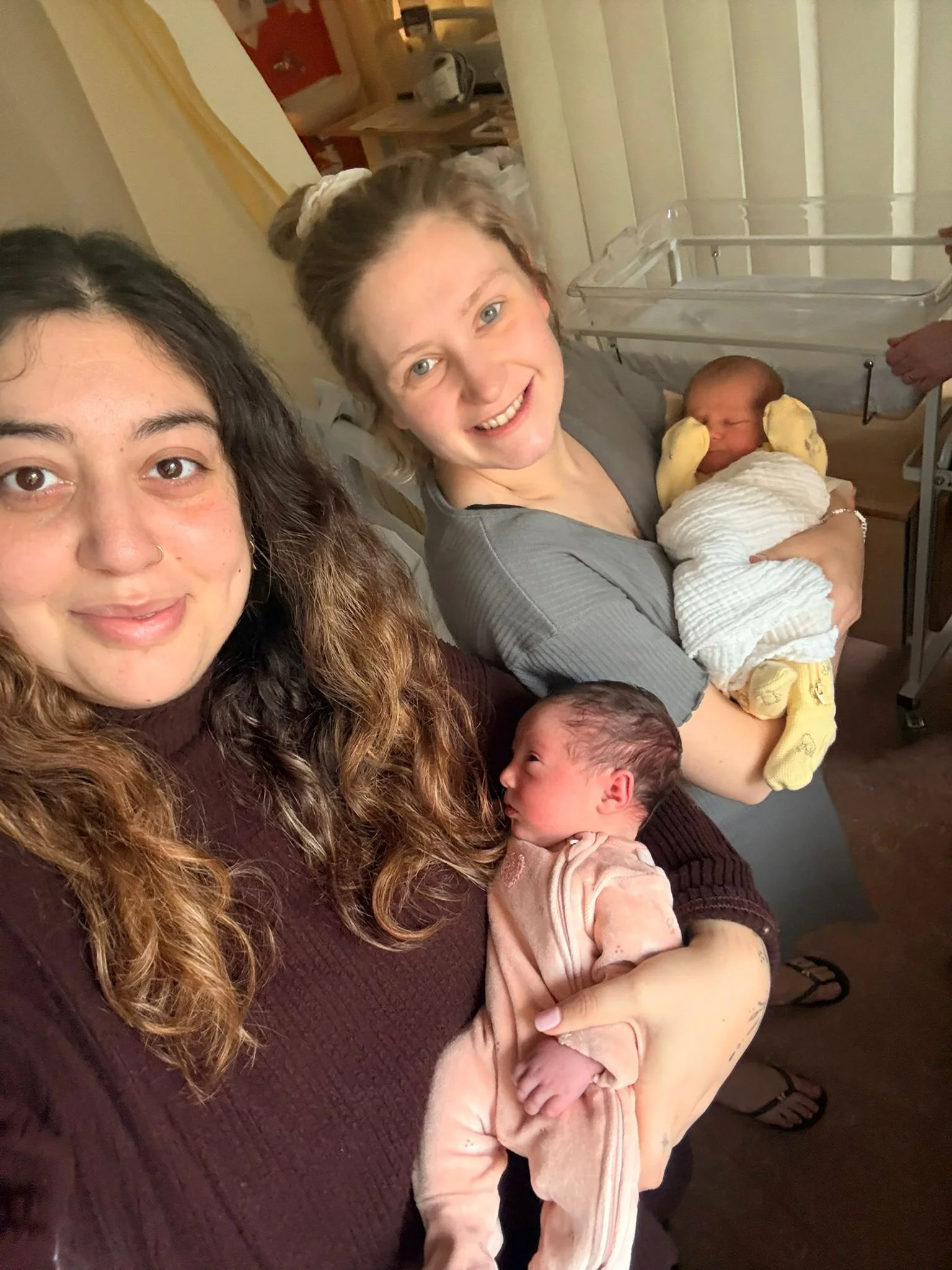 Two women holding newborn babies, smiling at the camera in a cozy indoor setting.