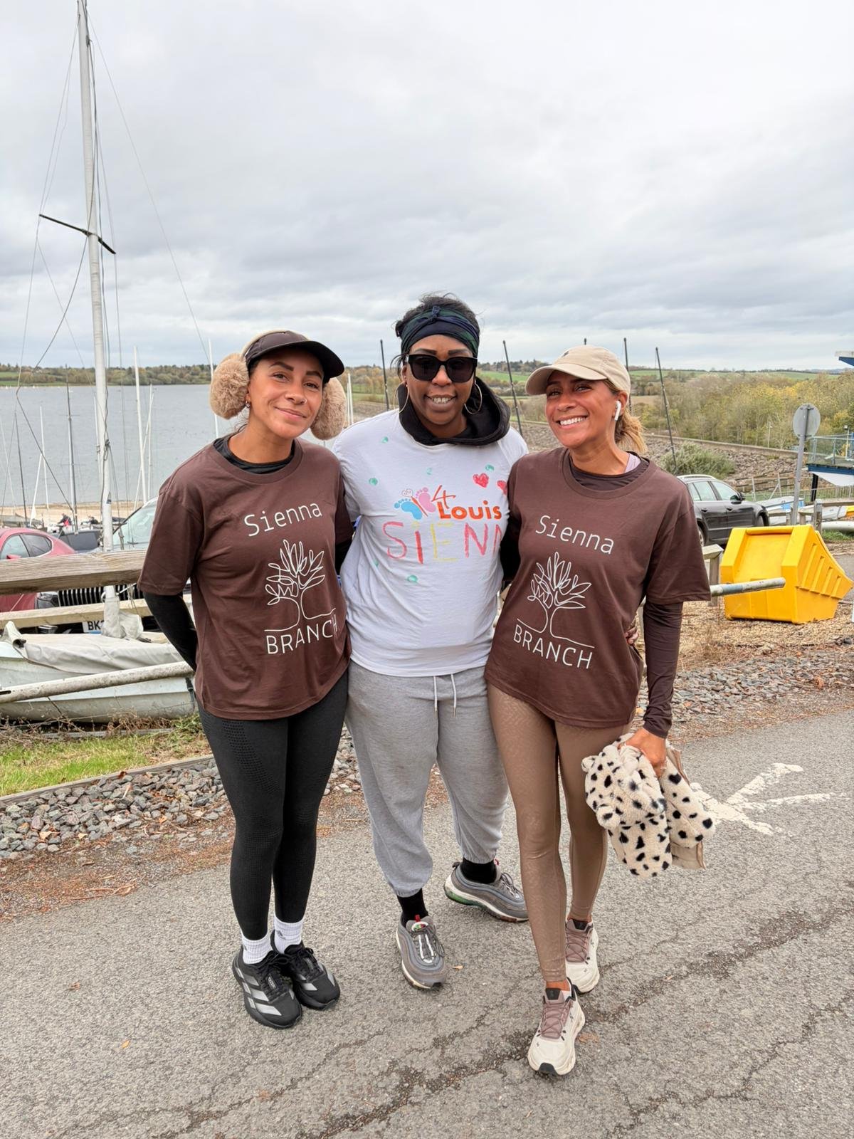 Three women standing outdoors by a body of water on a cloudy day. They are smiling and wearing casual athletic clothing, with two of them in matching brown shirts that say "Sienna Branch" and the third in a white shirt with colorful writing.