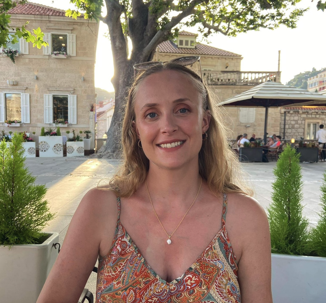 Smiling woman with wavy blonde hair, wearing sunglasses on her head, a patterned dress, a pearl necklace, and earrings, sitting outdoors at a restaurant or cafe with other people, trees, and historic buildings in the background.