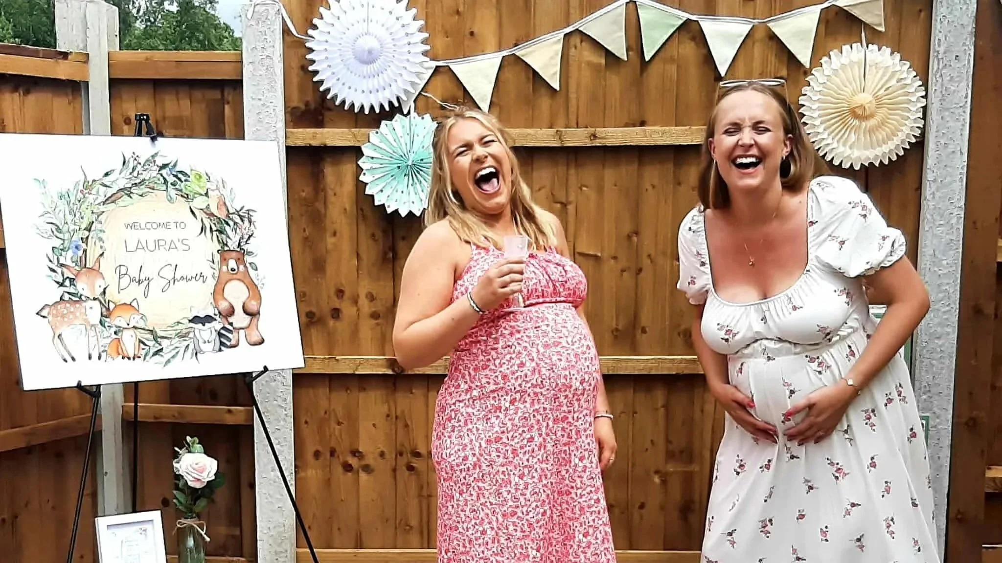 Two women laughing and posing at a baby shower, with a decorated wooden backdrop and a sign that says 'Welcome to Laura's Baby Shower' featuring animals and greenery.