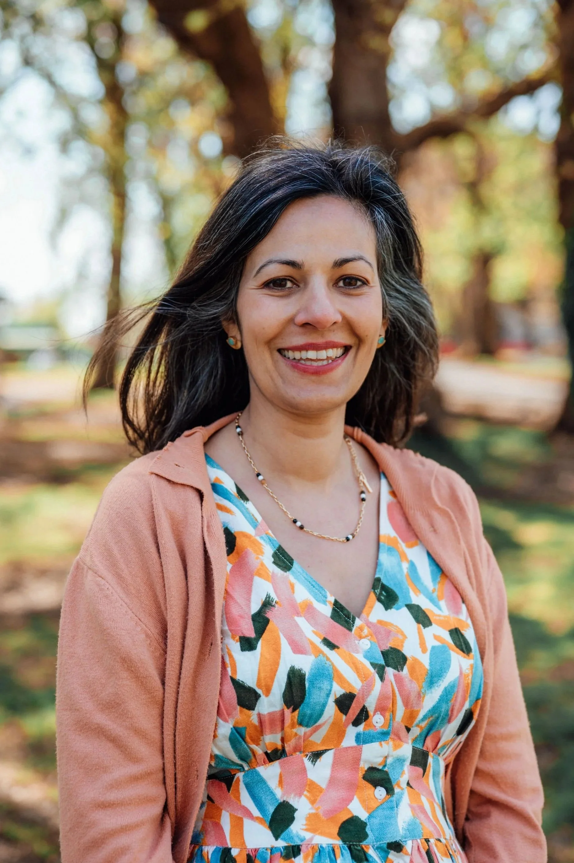 A woman with black hair and a smile standing outdoors in front of trees with colorful foliage, wearing a patterned dress and an orange cardigan.
