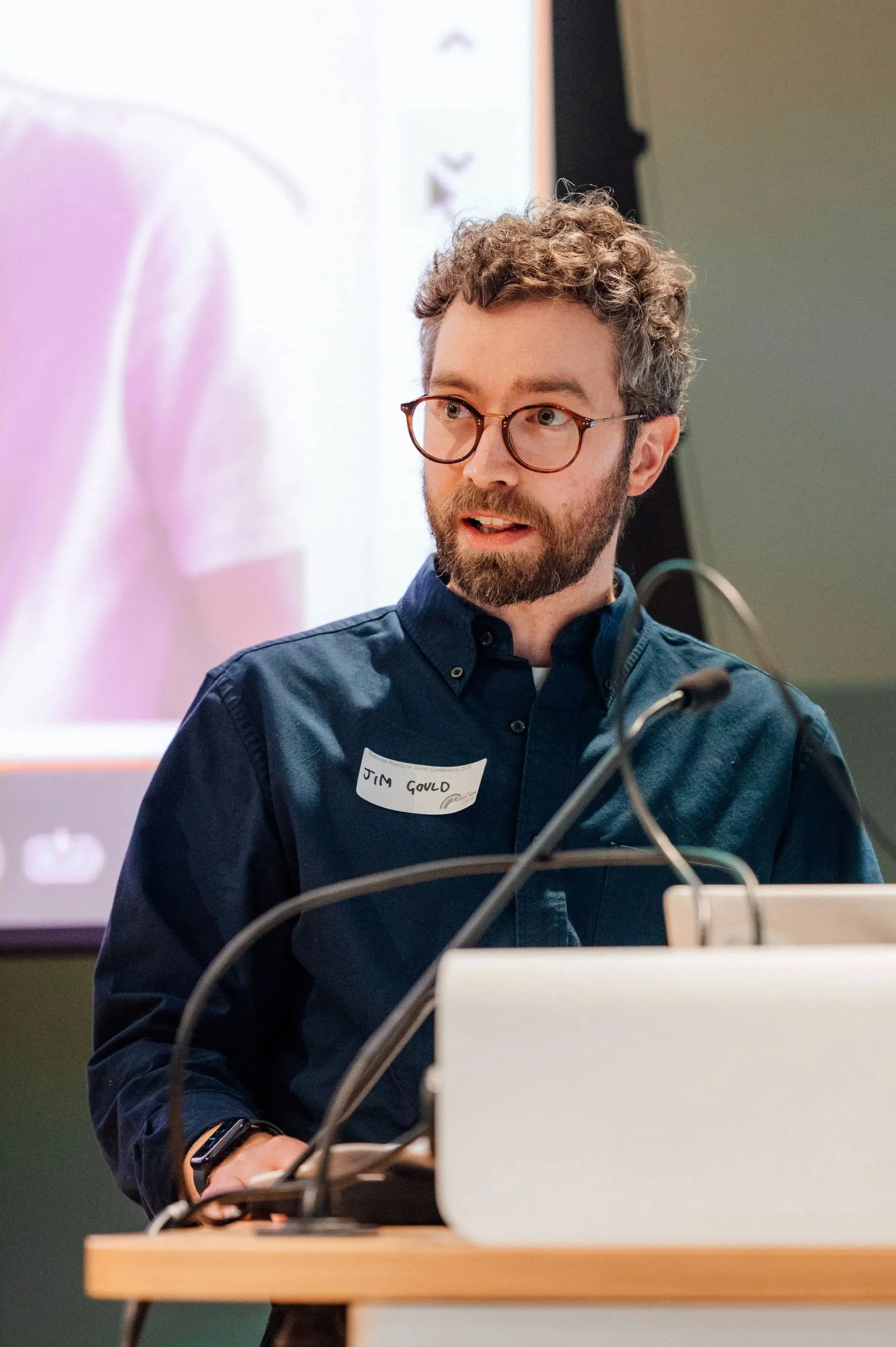 A man with curly hair and glasses, wearing a dark blue shirt with a name tag that says 'Jim Gould', standing at a podium in front of a large screen, giving a presentation or speech.