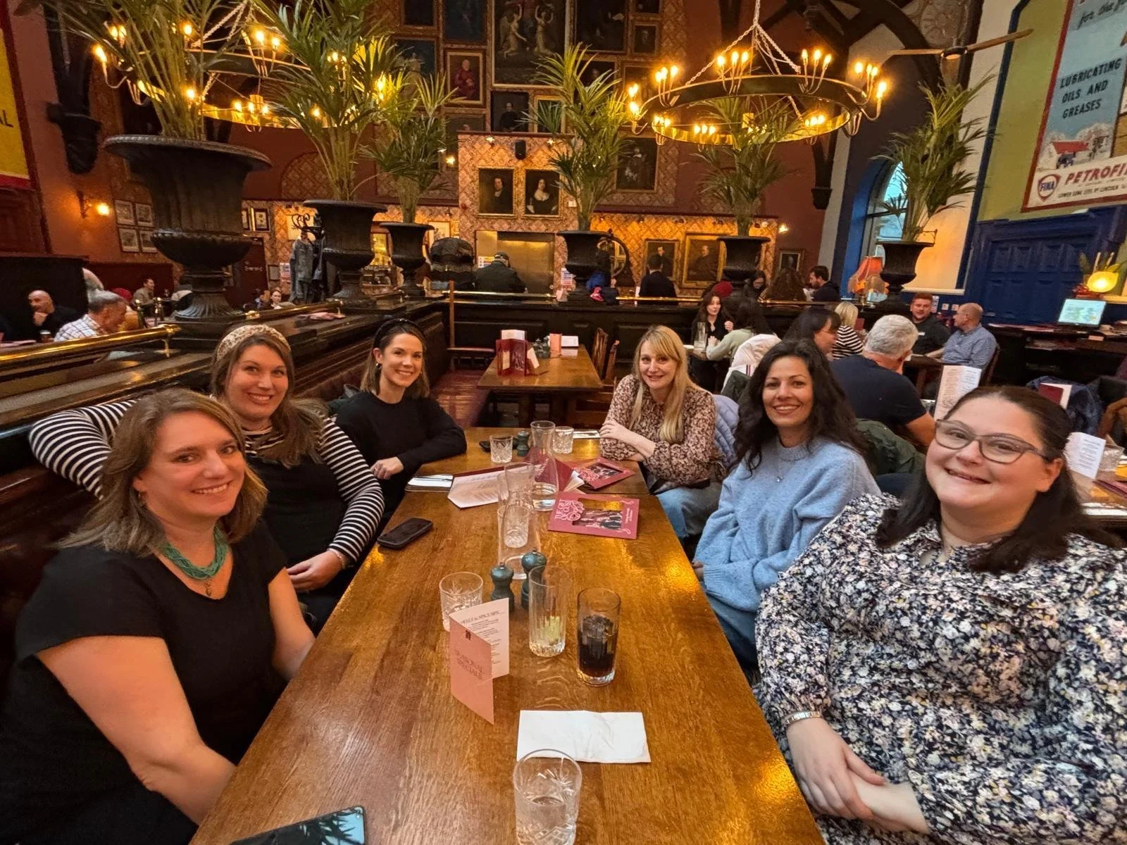 A group of six women sitting at a long wooden table in a warmly lit restaurant, smiling at the camera. The background features ornate decor with framed pictures, large black planters with greenery, and warm ambient lighting.