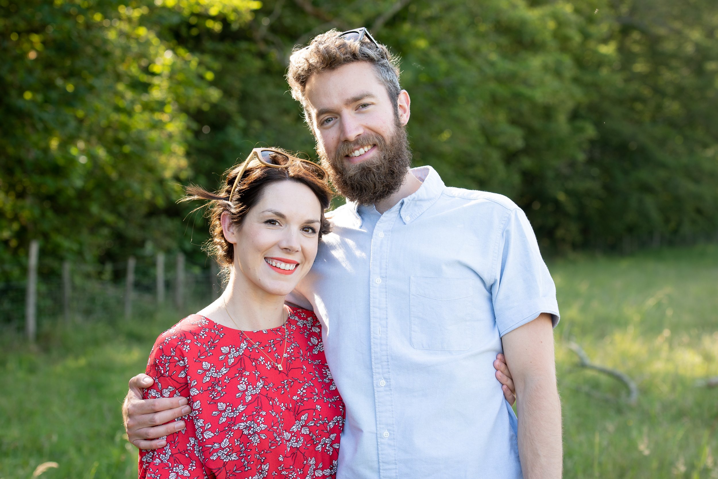 A smiling man with a beard in a light blue shirt standing outdoors with a woman in a red floral dress who is also smiling. They are embracing with greenery in the background.
