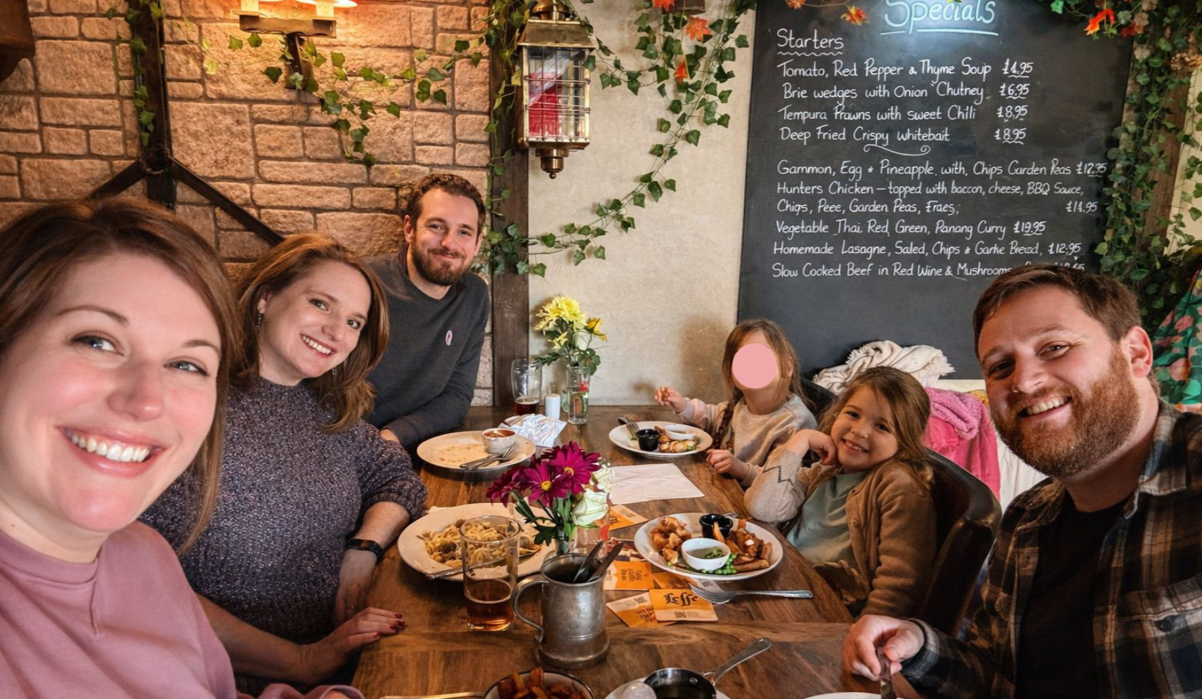 A group of six people, including three adults and three children, sitting at a restaurant table for a meal, with a blackboard menu in the background and a brick wall with greenery.
