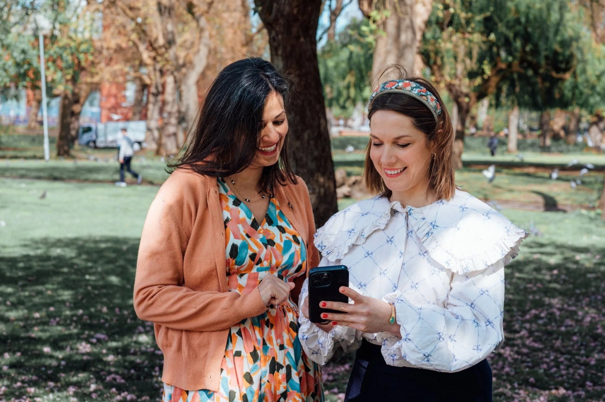 Two women standing in a park on a sunny day, looking at a smartphone together and smiling, with trees and other park visitors in the background.
