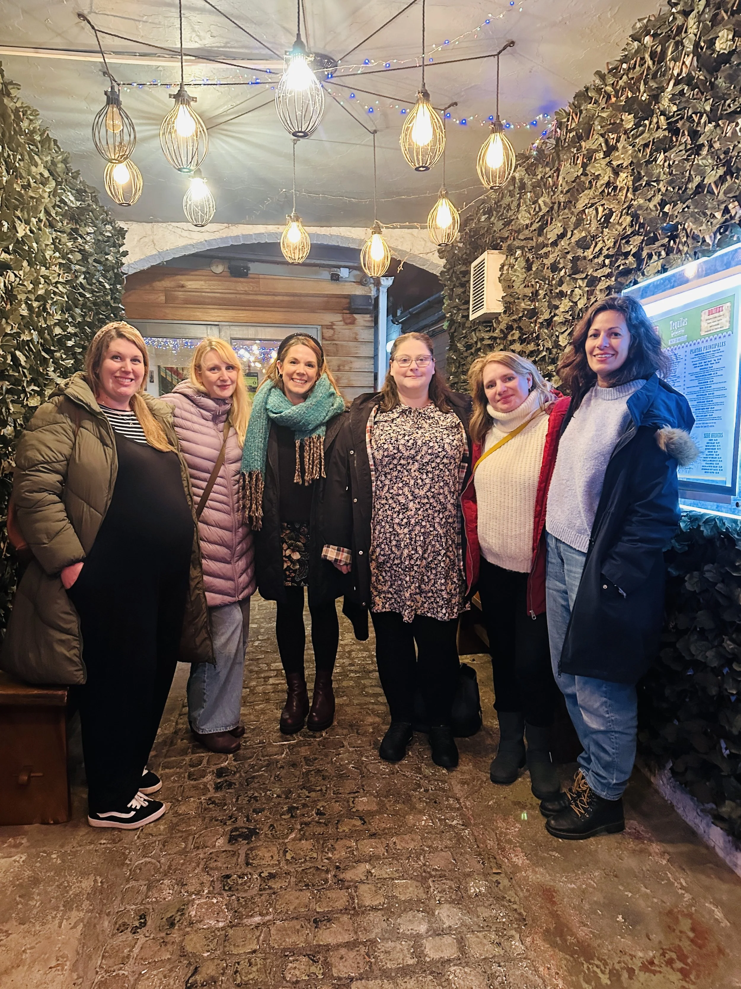 Six women standing together in an indoor space with warm lighting, cobblestone floor, and decorative greenery on the walls, smiling at the camera.
