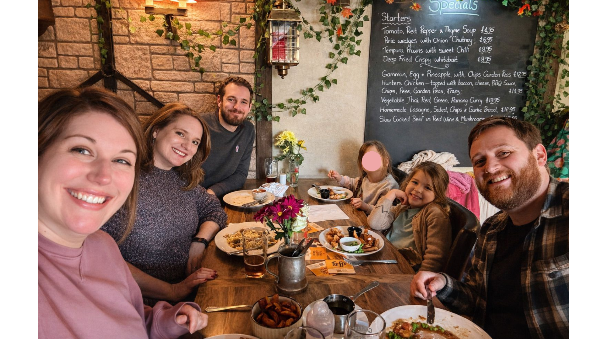 Family of six enjoying a meal at a restaurant, with a chalkboard menu on the wall behind them.
