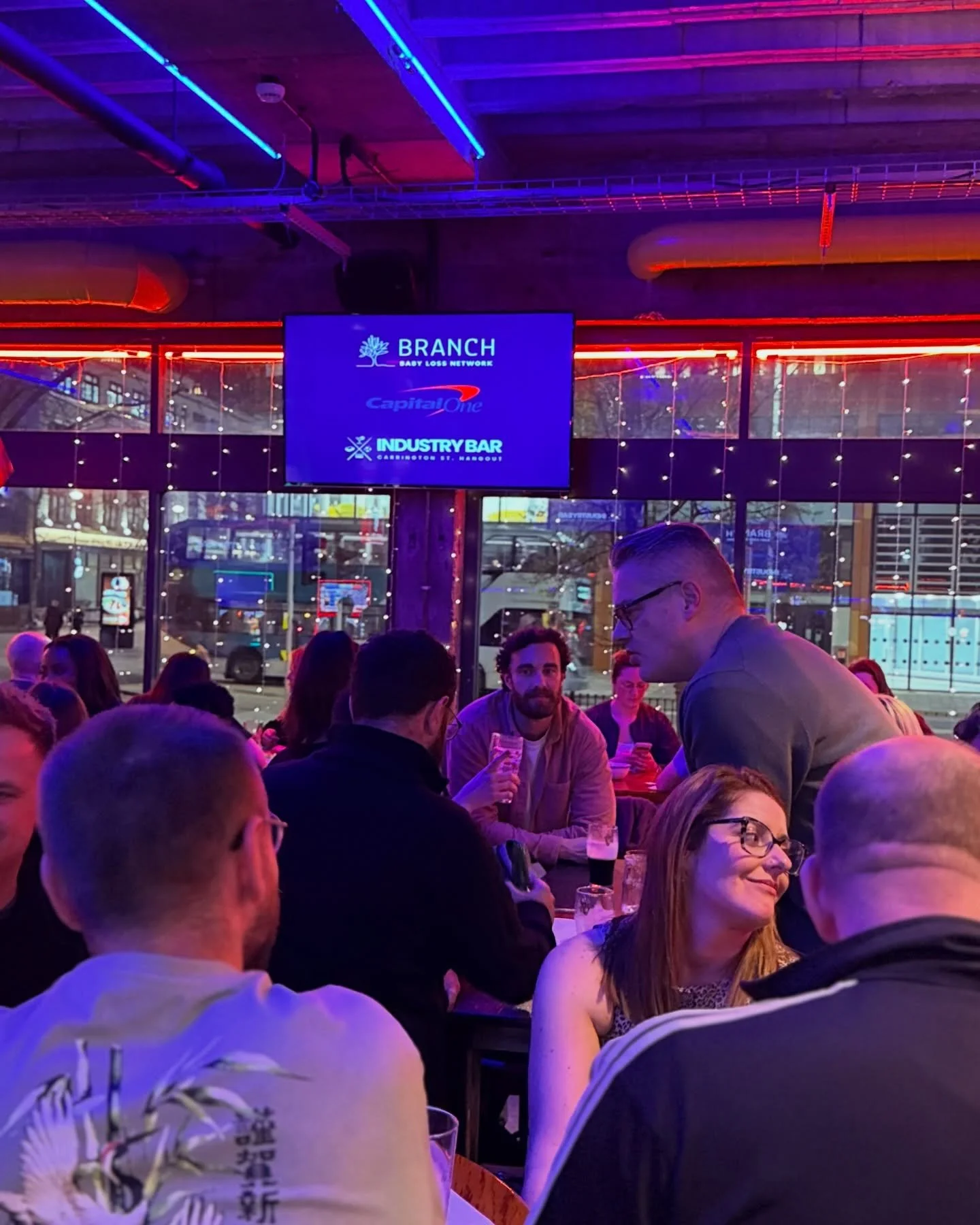 People dining at a bar or restaurant with neon lights, and a large screen displaying the words 'BRANCH Baby Loss Network,' 'Capital One,' and 'Industry Bar'.