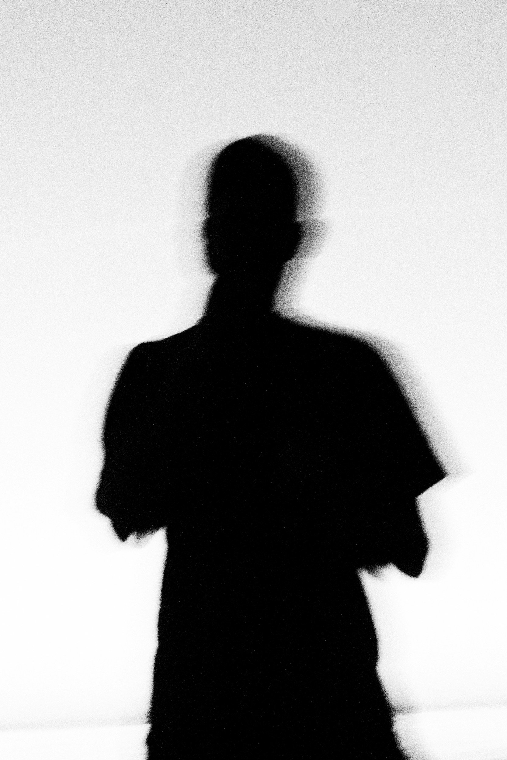 A silhouette of a person with short hair, standing against a light background, with a blurry and grainy appearance.