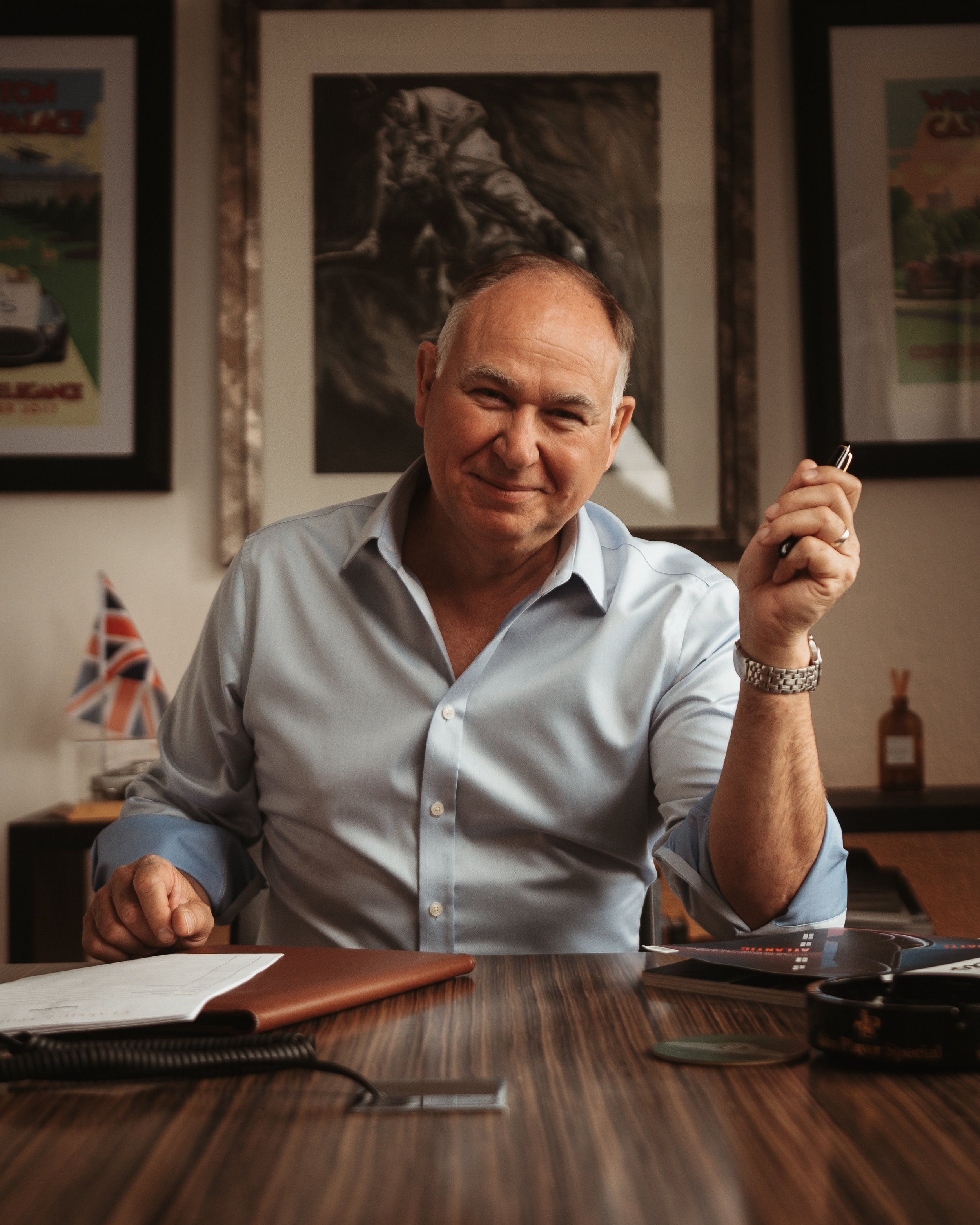 A middle-aged man with a light shirt and watch, sitting at a wooden desk, smiling, holding a pen, with a notebook and magazines on the desk, in a room with framed artwork on the wall.