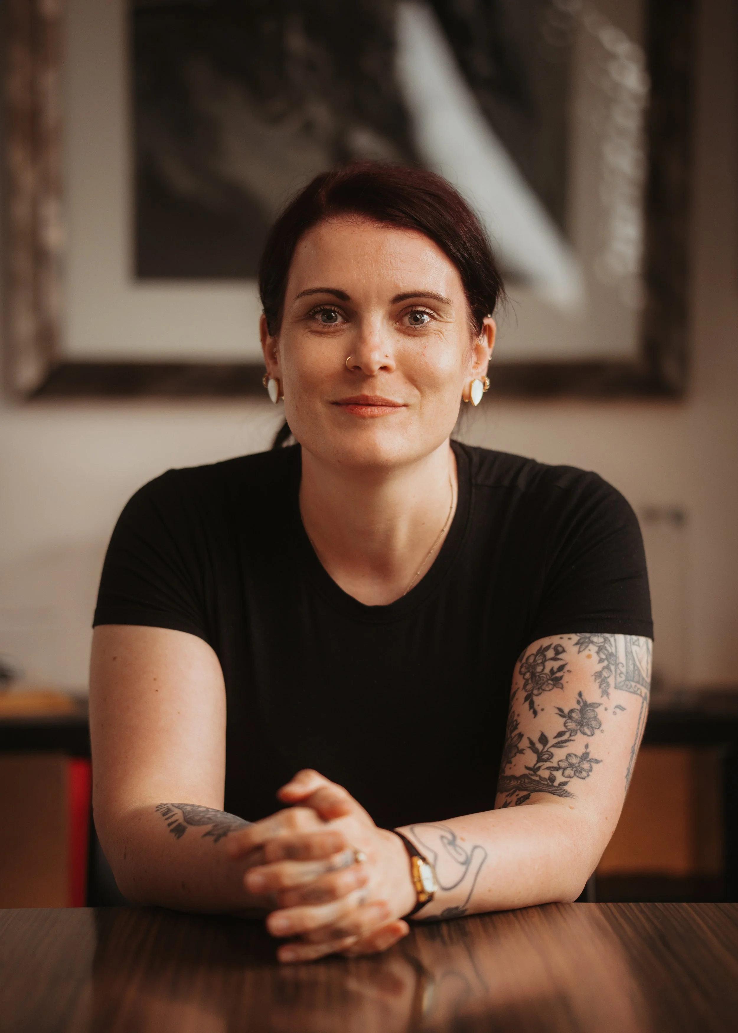 A woman with short dark hair and tattoos on her arms, sitting at a wooden table with her hands clasped, wearing a black shirt and earrings, in a warmly lit room with framed artwork in the background.