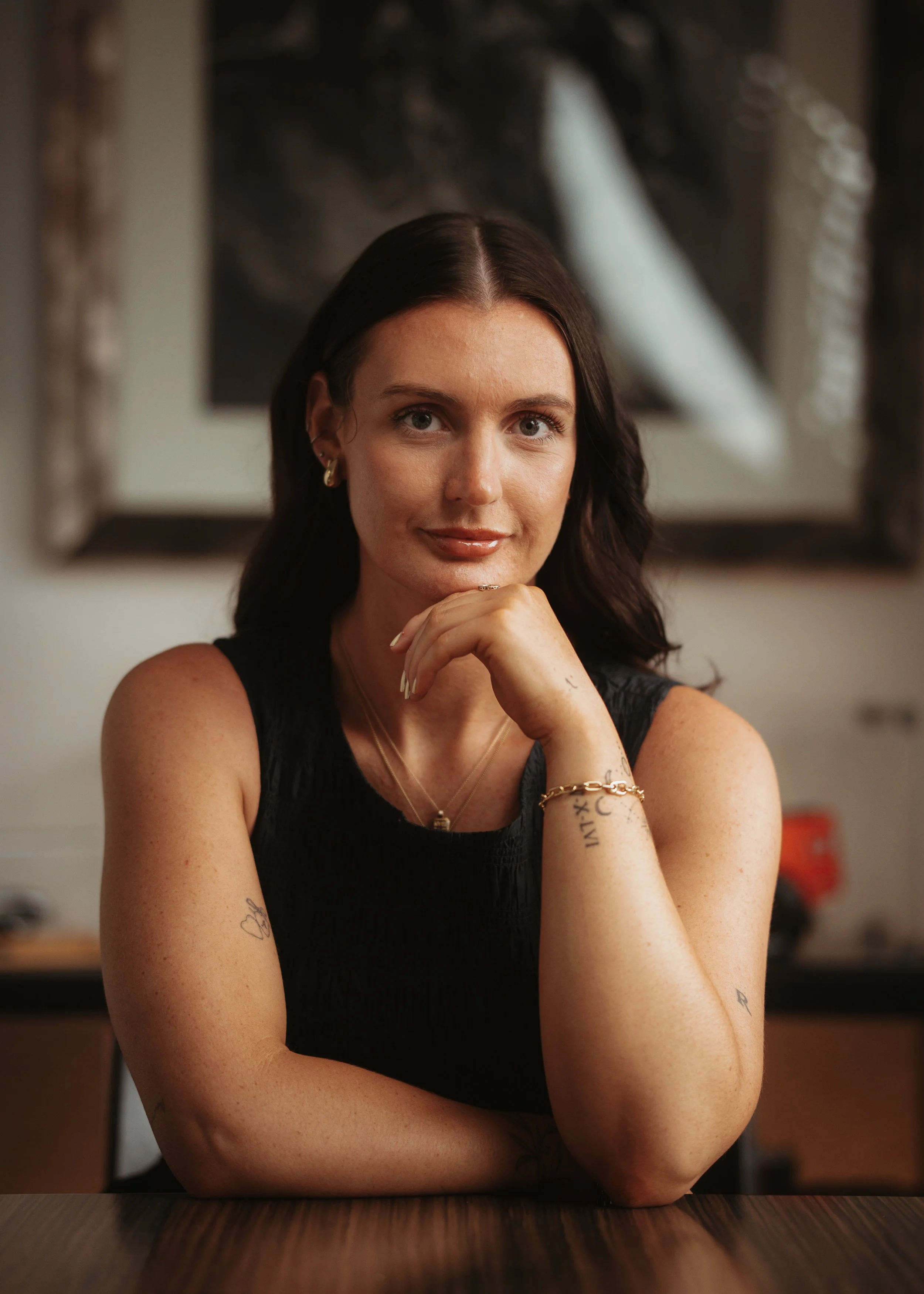 A woman with dark hair and tattoos on her arms, sitting at a table, resting her chin on her hand, looking at the camera with a slight smile. She is wearing earrings, layered necklaces, a bracelet, and a sleeveless dark top. The background is blurred with a framed picture or artwork behind her.