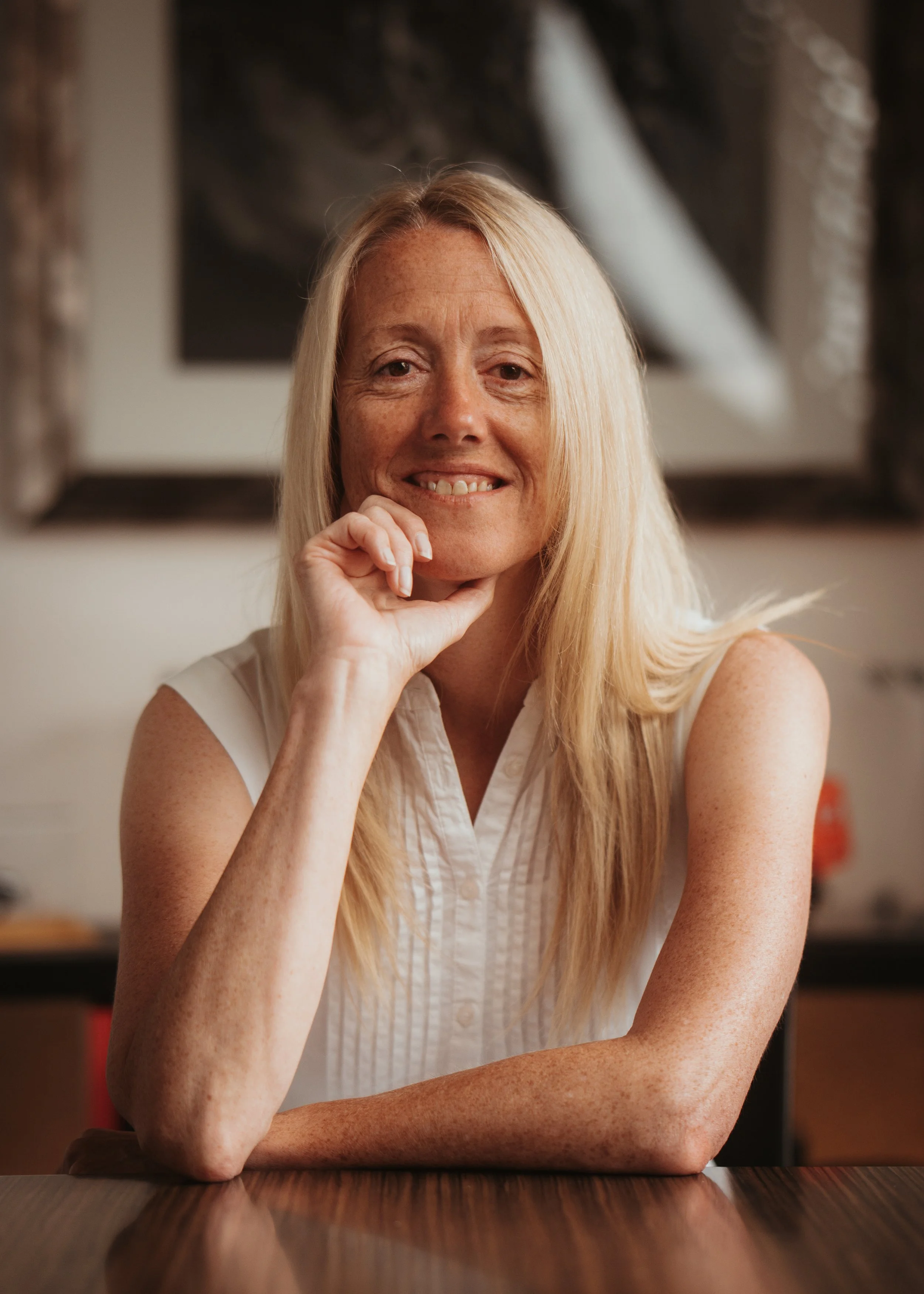 Portrait of a blonde woman smiling with her chin resting on her hand, sitting at a wooden table in a room with framed art on the wall in the background.