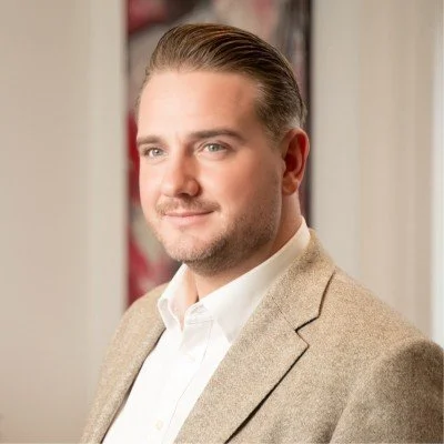 A man with neatly combed brown hair, wearing a white shirt and a beige blazer, standing indoors with a neutral expression.