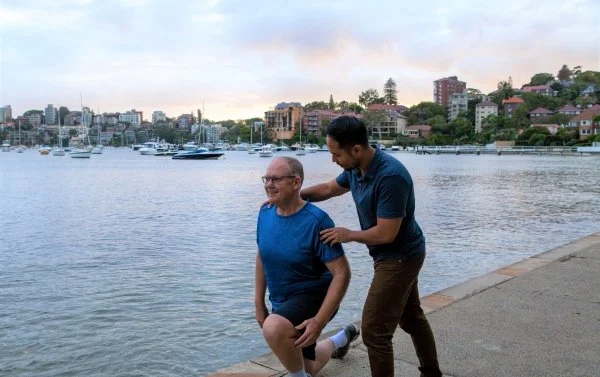 Personal trainer guiding an older adult through a lunge during an outdoor training session by the water.